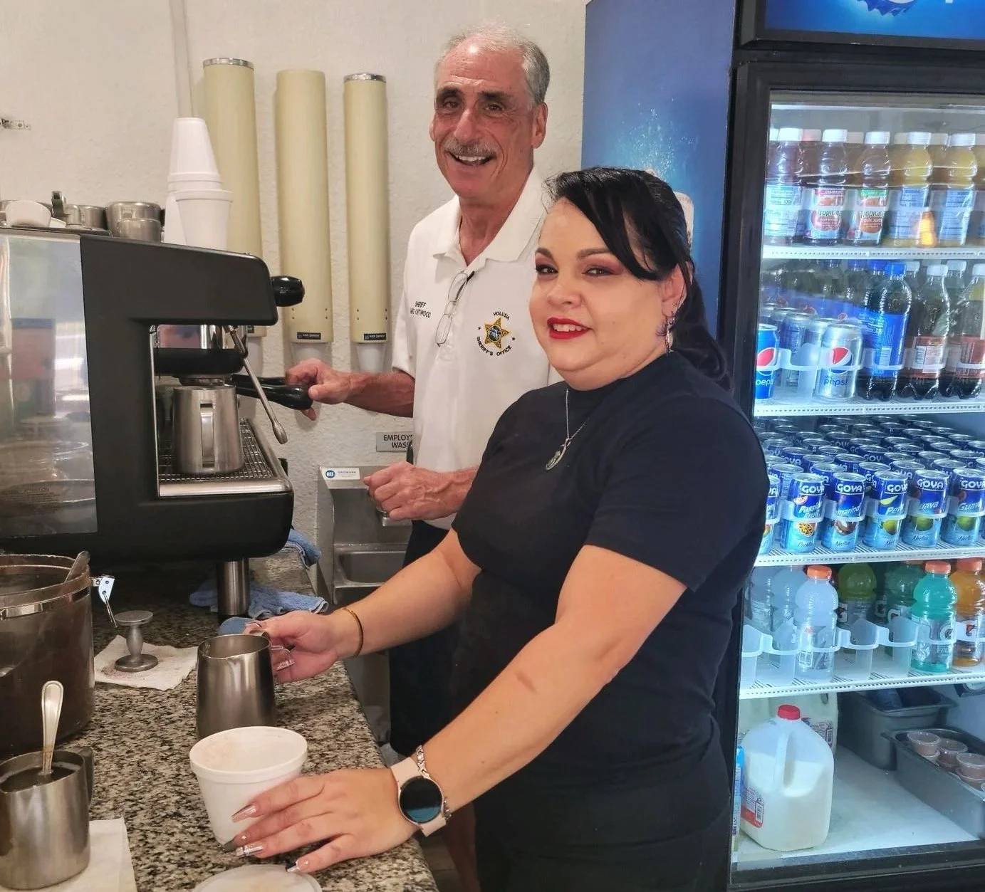 A man and woman in a cafe, the man is operating an espresso machine and the woman is pouring a beverage into a cup, with a refrigerator filled with drinks in the background.