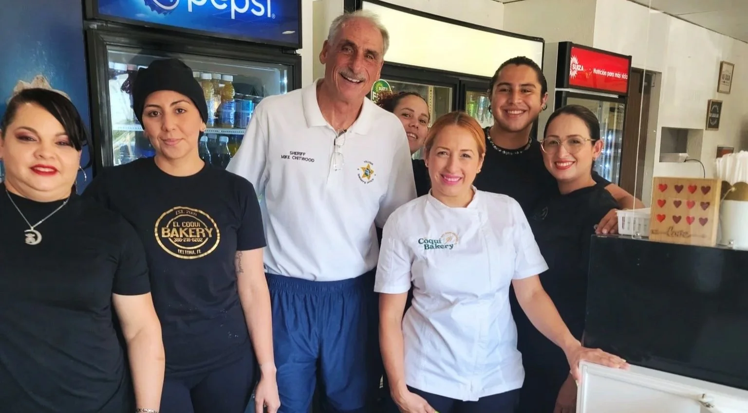Group of seven people, including a man in a sheriff's uniform, posing inside a bakery or cafe with refrigerators in the background.