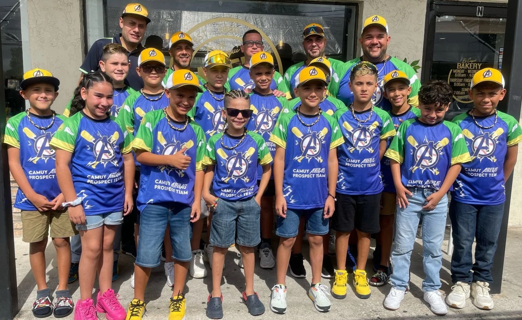 A group of children and young adults posing outdoors, wearing matching blue, green, and yellow jerseys and yellow caps, with some wearing sunglasses and chains, in front of a storefront.