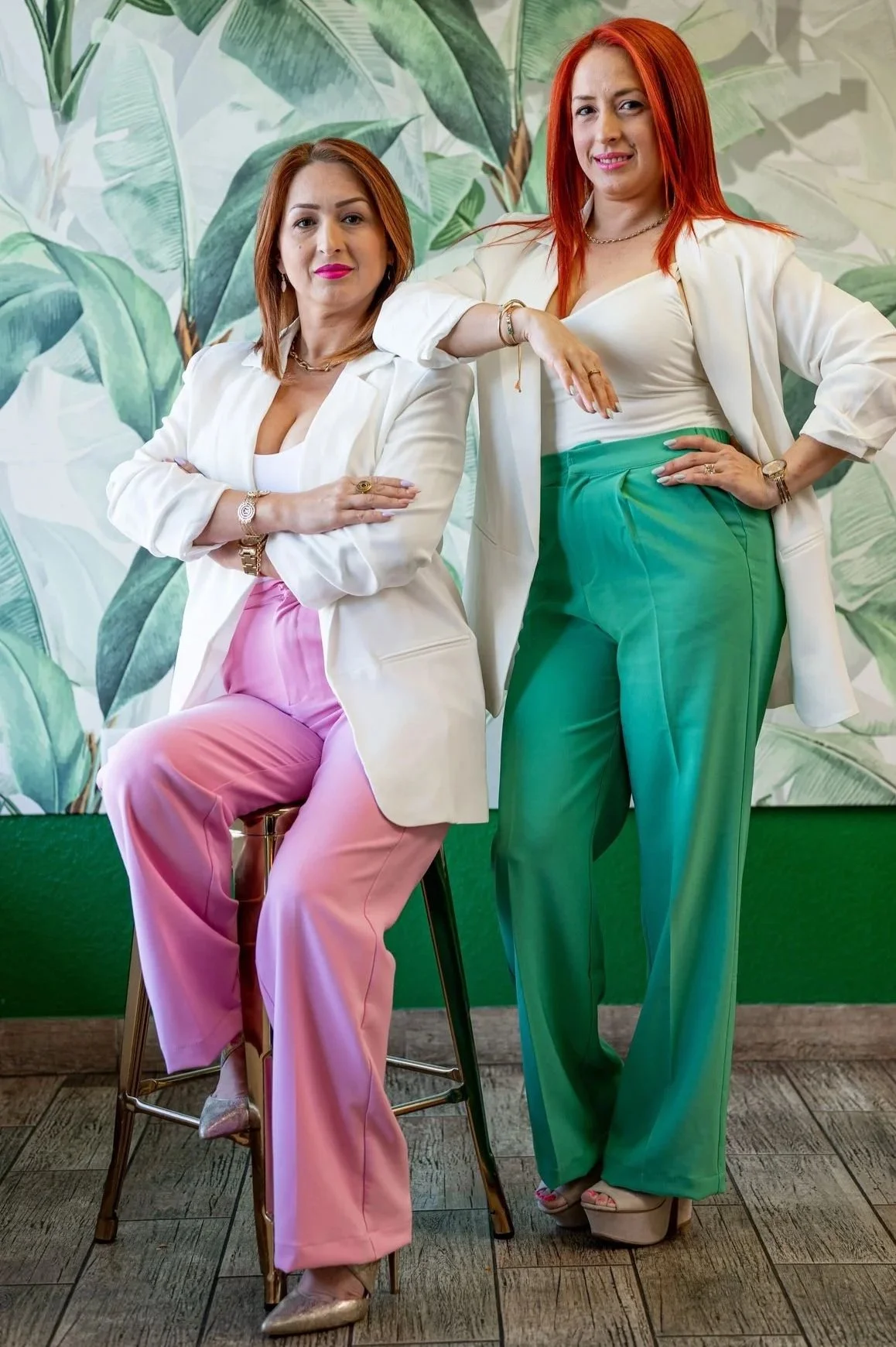 Two women with red hair pose confidently in front of a tropical leaf background. One is sitting on a stool, wearing a white blazer, pink wide-leg pants, and gold shoes. The other stands beside her with her arm resting on the sitting woman's shoulder,