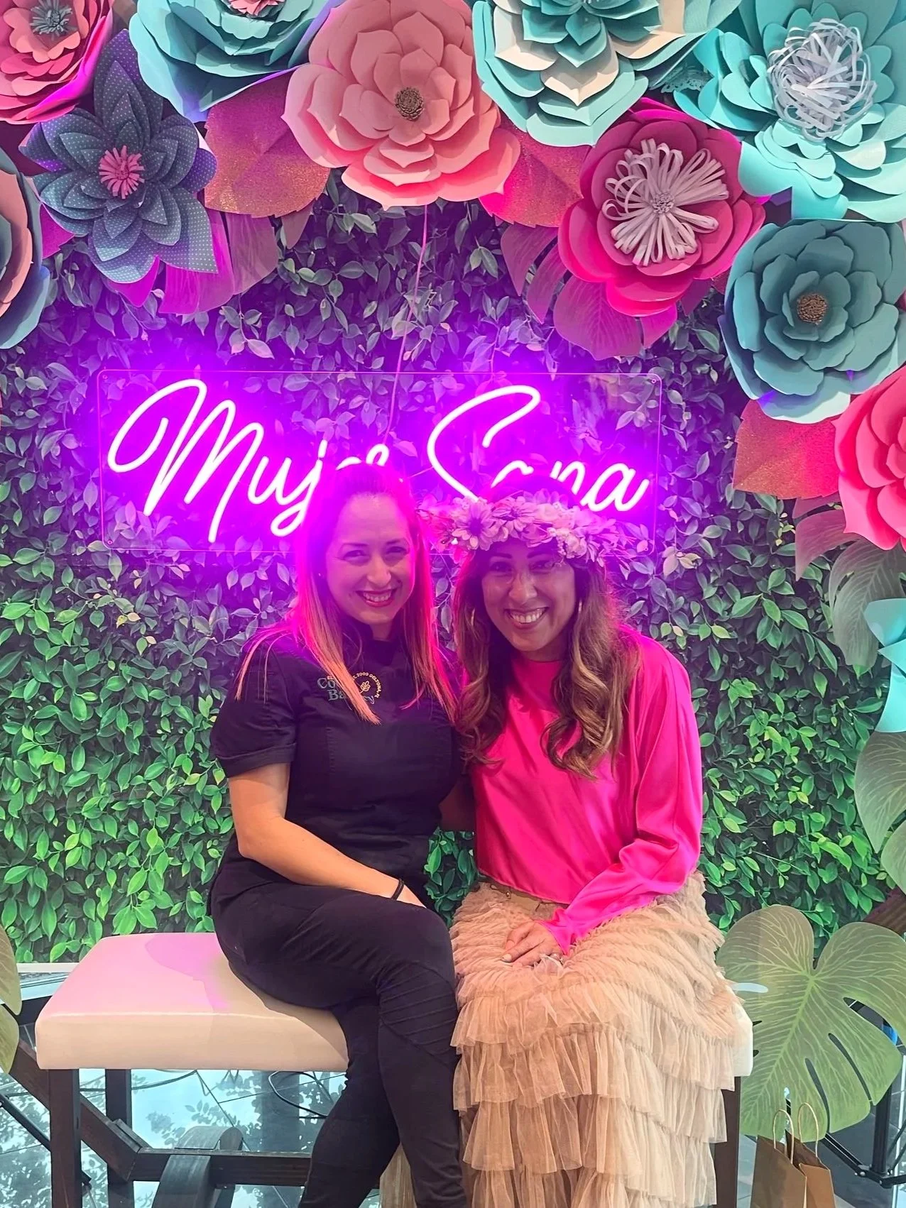 Two women sitting together, smiling at the camera, with a background of colorful paper flowers and a neon pink sign that says 'Mija Sana'. One woman is wearing a black uniform and the other is wearing a pink top and a floral crown.