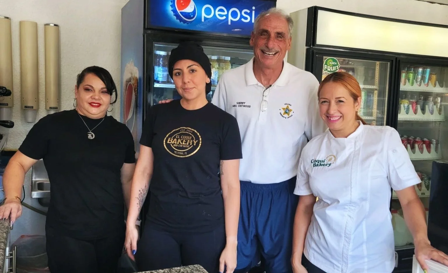 Four people standing behind a counter in a bakery, smiling at the camera. They are inside a store with a Pepsi sign and refrigerator in the background. The group includes two women in black and white bakery uniforms, a man in a sheriff's uniform, and