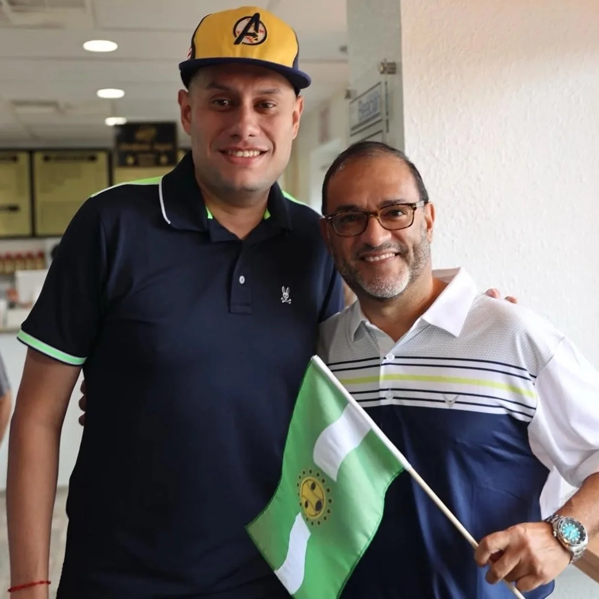 Two men standing indoors, one holding a green and white flag with a yellow emblem, smiling at the camera. One man is wearing a dark polo shirt and a yellow cap with a Marvel Avengers logo, and the other is wearing glasses and a white and blue striped