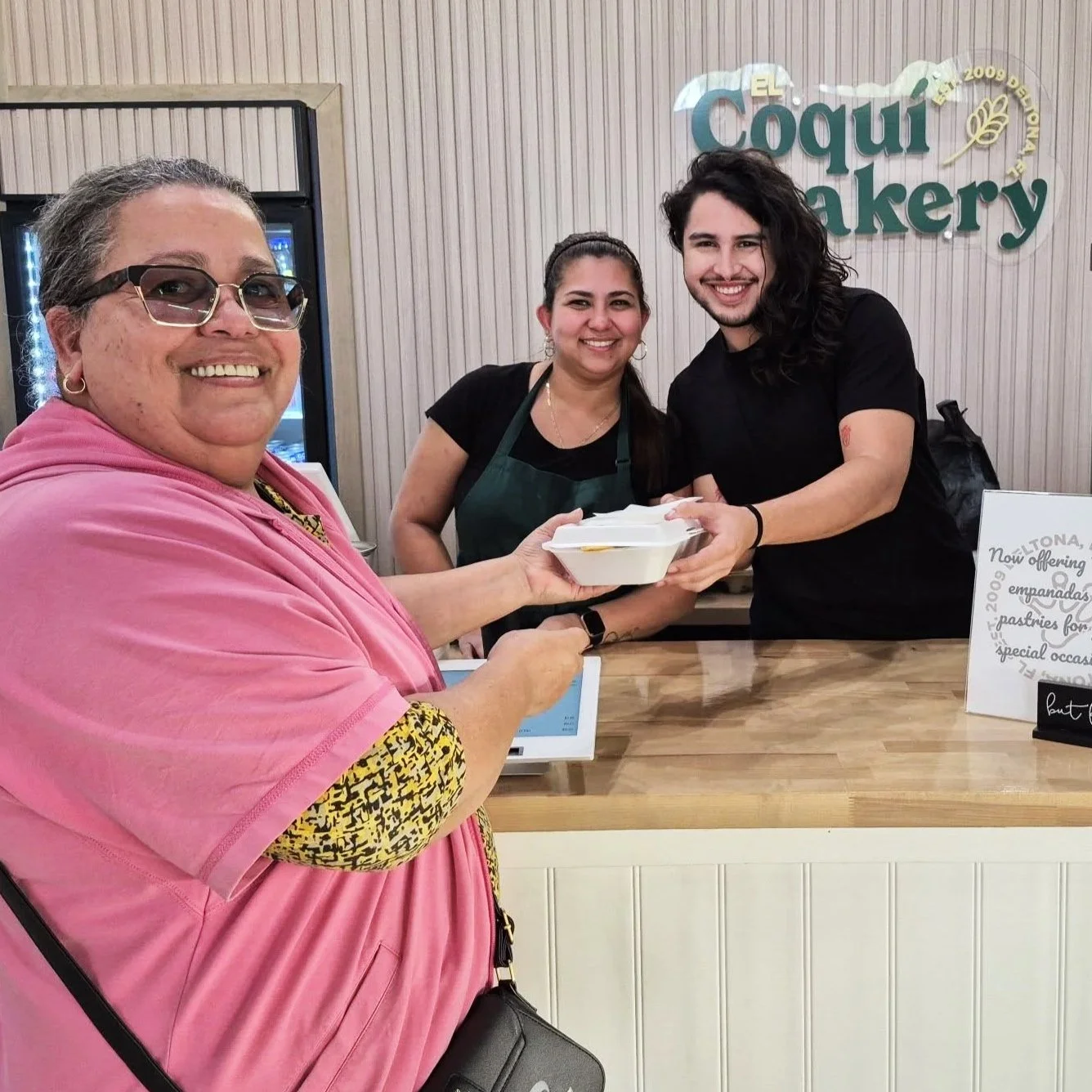 Three women smiling at a bakery counter, with one woman handing a takeout container to another. The bakery sign in the background reads 'Coqui Bakery'.