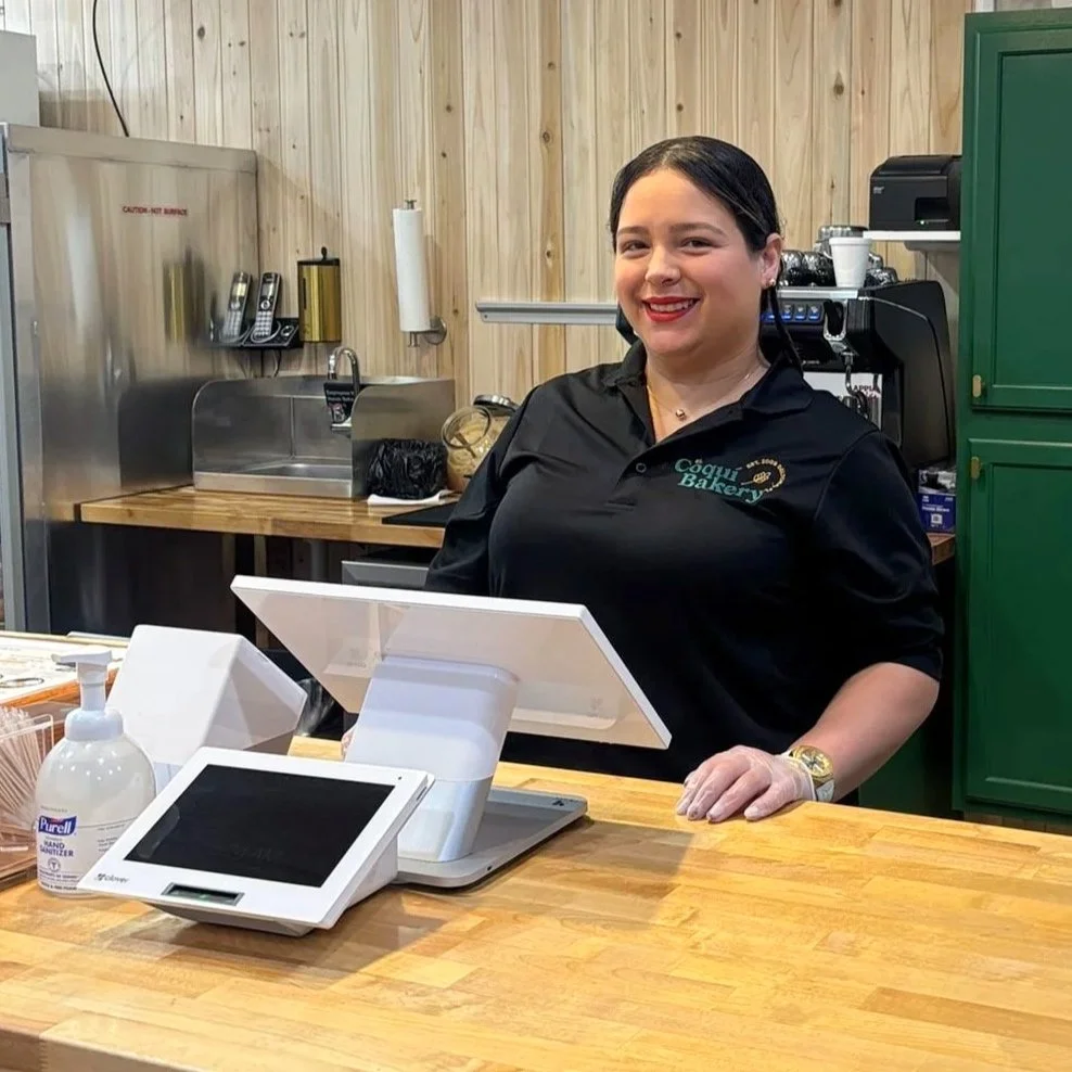 A woman with dark hair smiling behind a bakery counter, wearing a black shirt with a bakery logo on it.