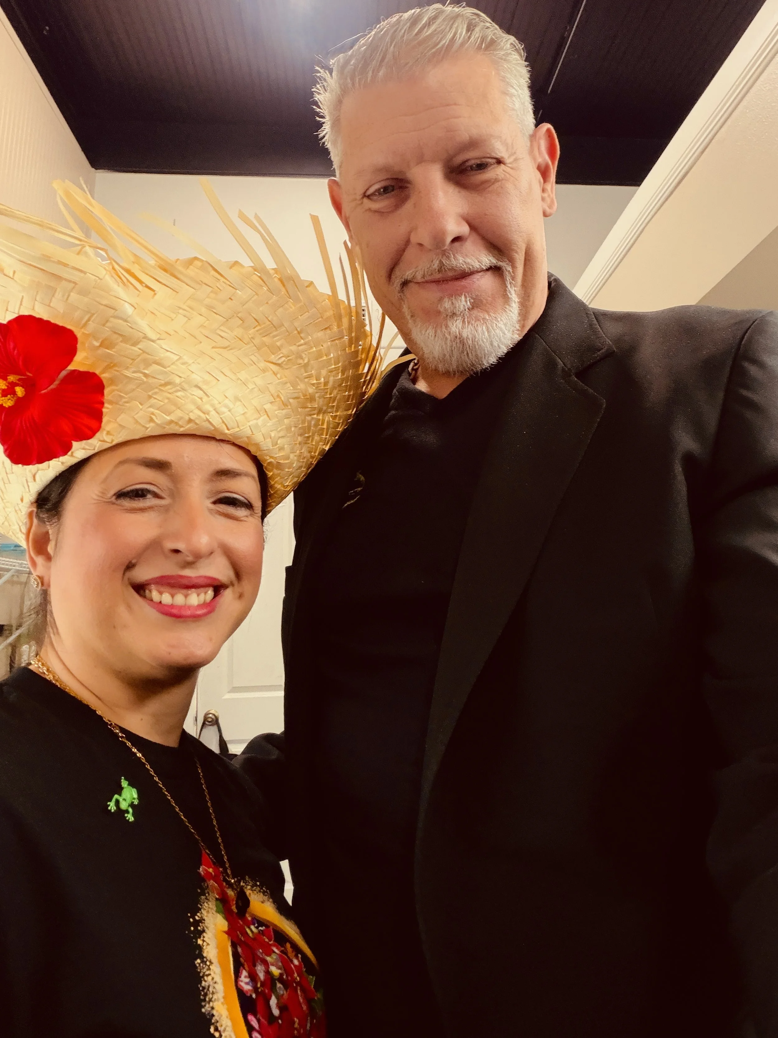 A woman smiling and wearing a large straw hat with a red flower and a woman with gray hair and a beard standing indoors.
