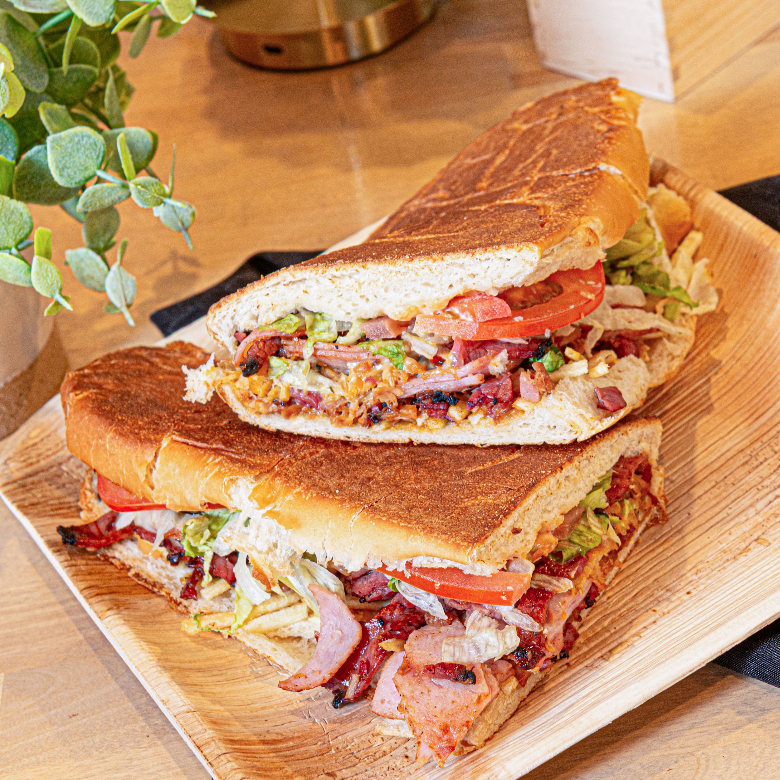 Two halves of a toasted sub sandwich with visible layers of bacon, lettuce, tomato, and meat, placed on a wooden tray.