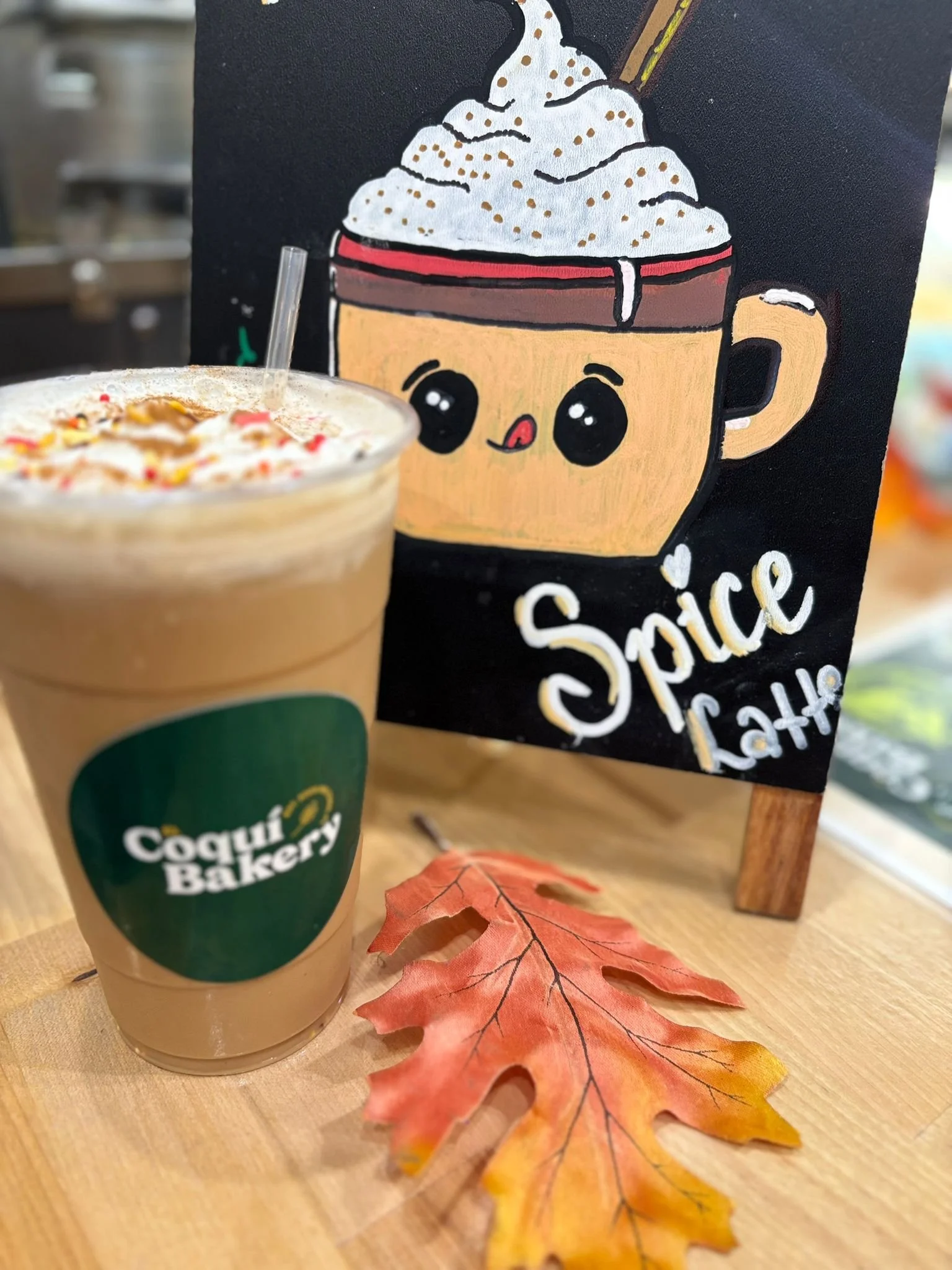 A mocha latte with whipped cream and colorful sprinkles on top, placed on a wooden table. Next to the drink is a drying fall leaf. Behind is a black chalkboard sign with a cute cartoon coffee cup with whipped cream and a straw, labeled 'Spice Latte'.