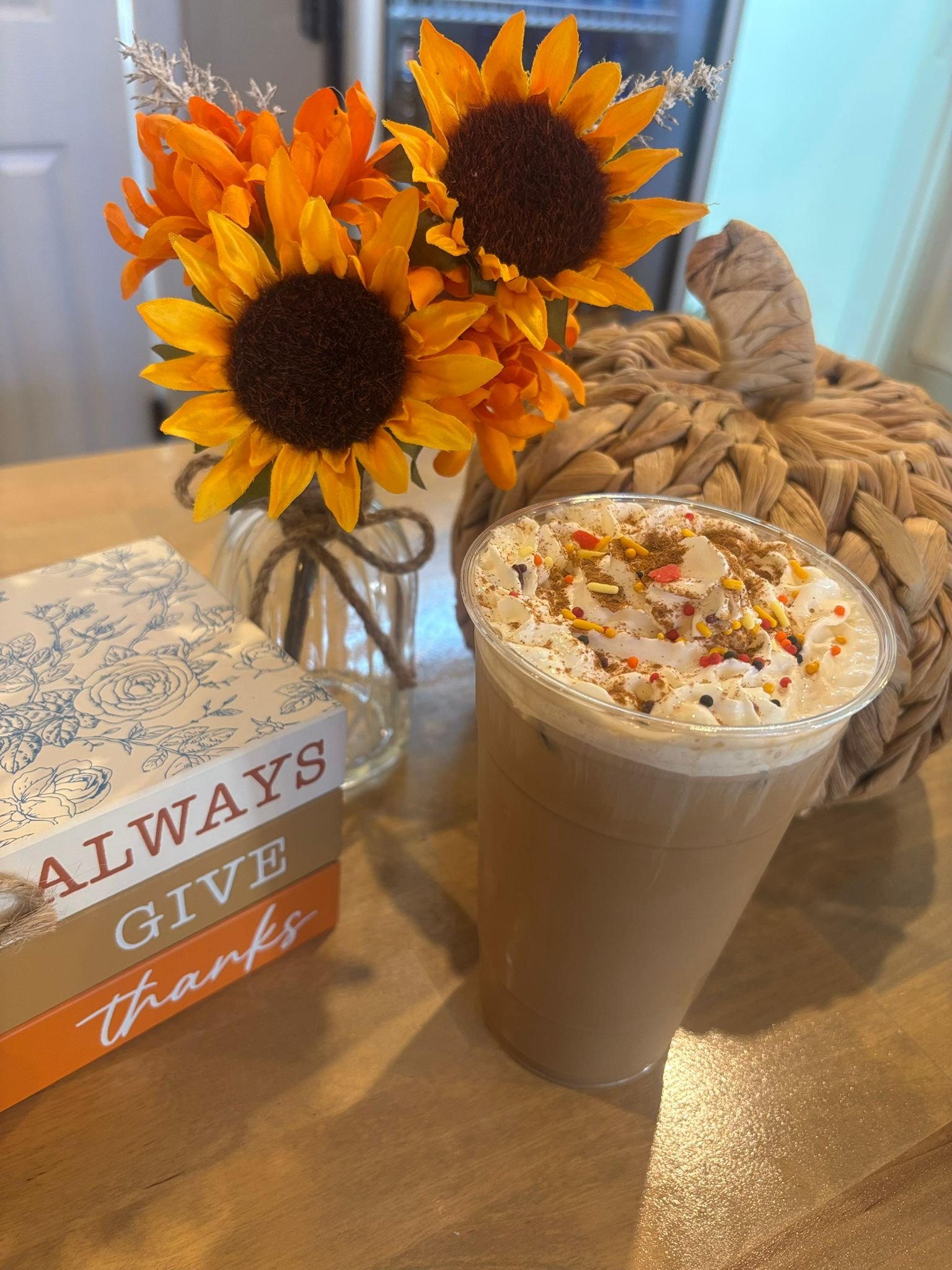 A glass of chocolate milkshake topped with whipped cream, sprinkles, and chocolate shavings on a wooden table next to a bouquet of sunflower flowers in a jar, a decorative pumpkin, and a box with the text 'Always Give Thanks'.