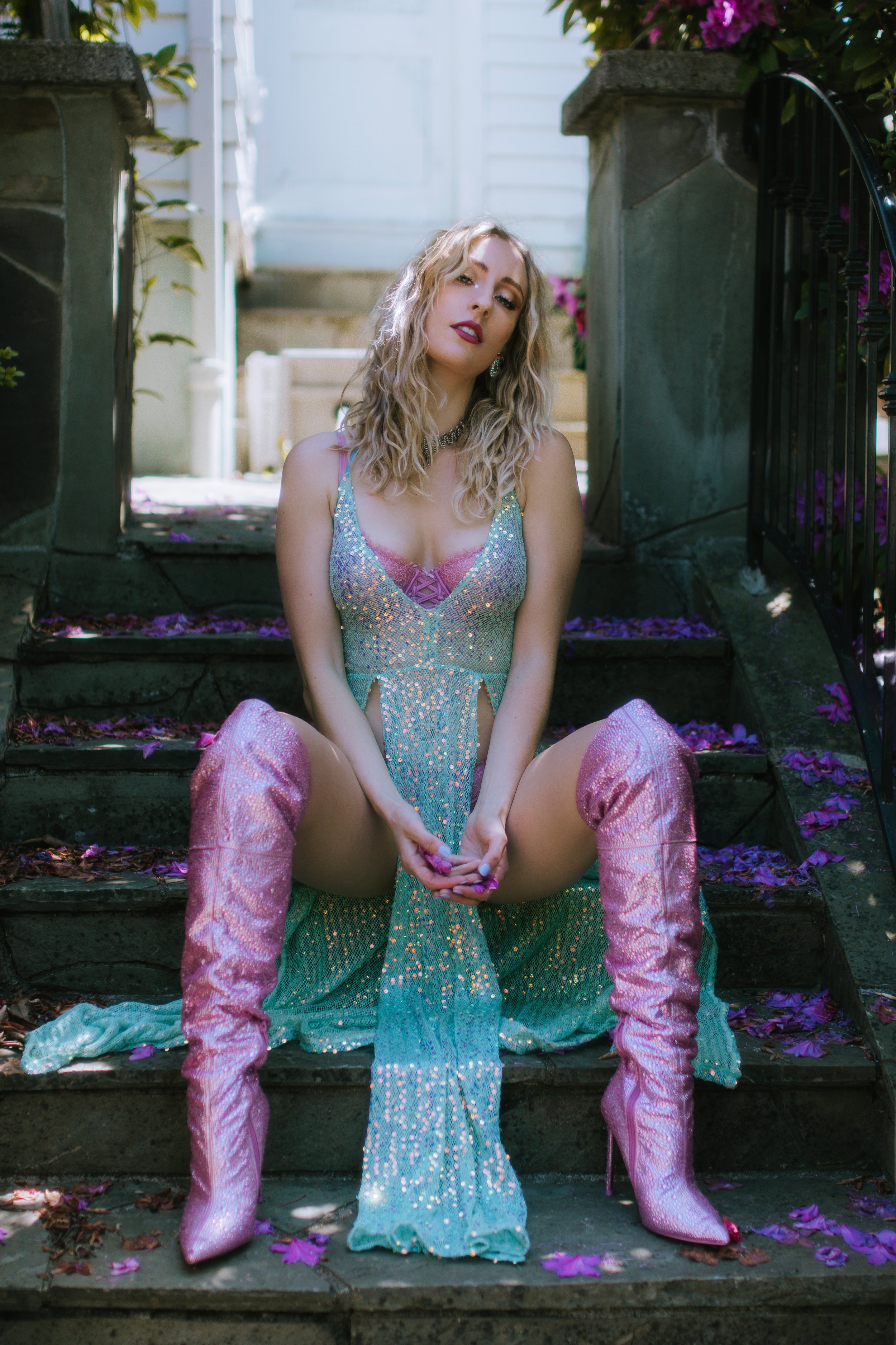 Woman in sparkly dress and pink boots sitting on steps outdoors with flowers around her.