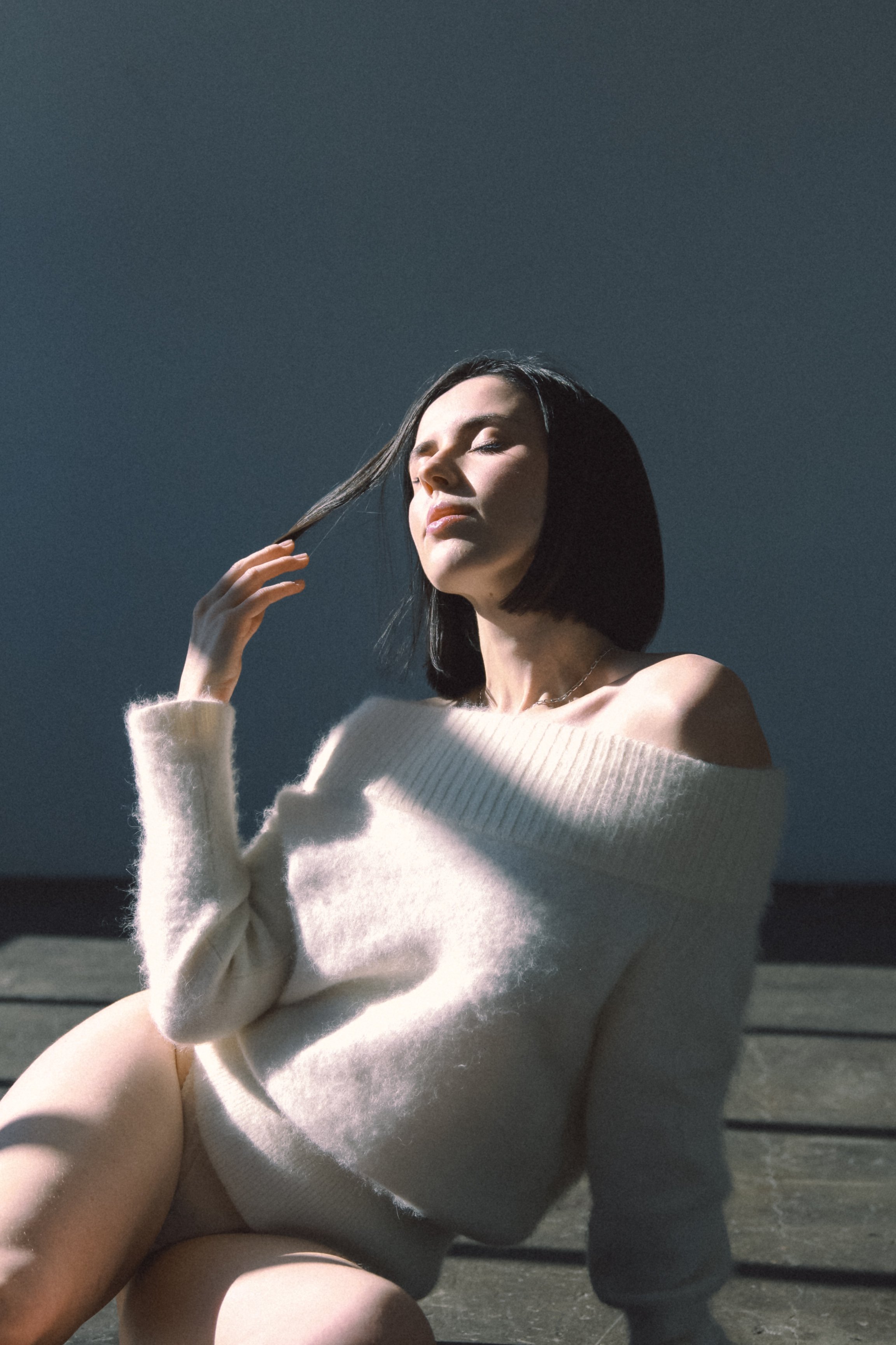 Woman in an off-shoulder sweater sitting on the floor, touching her hair, with eyes closed in soft sunlight.