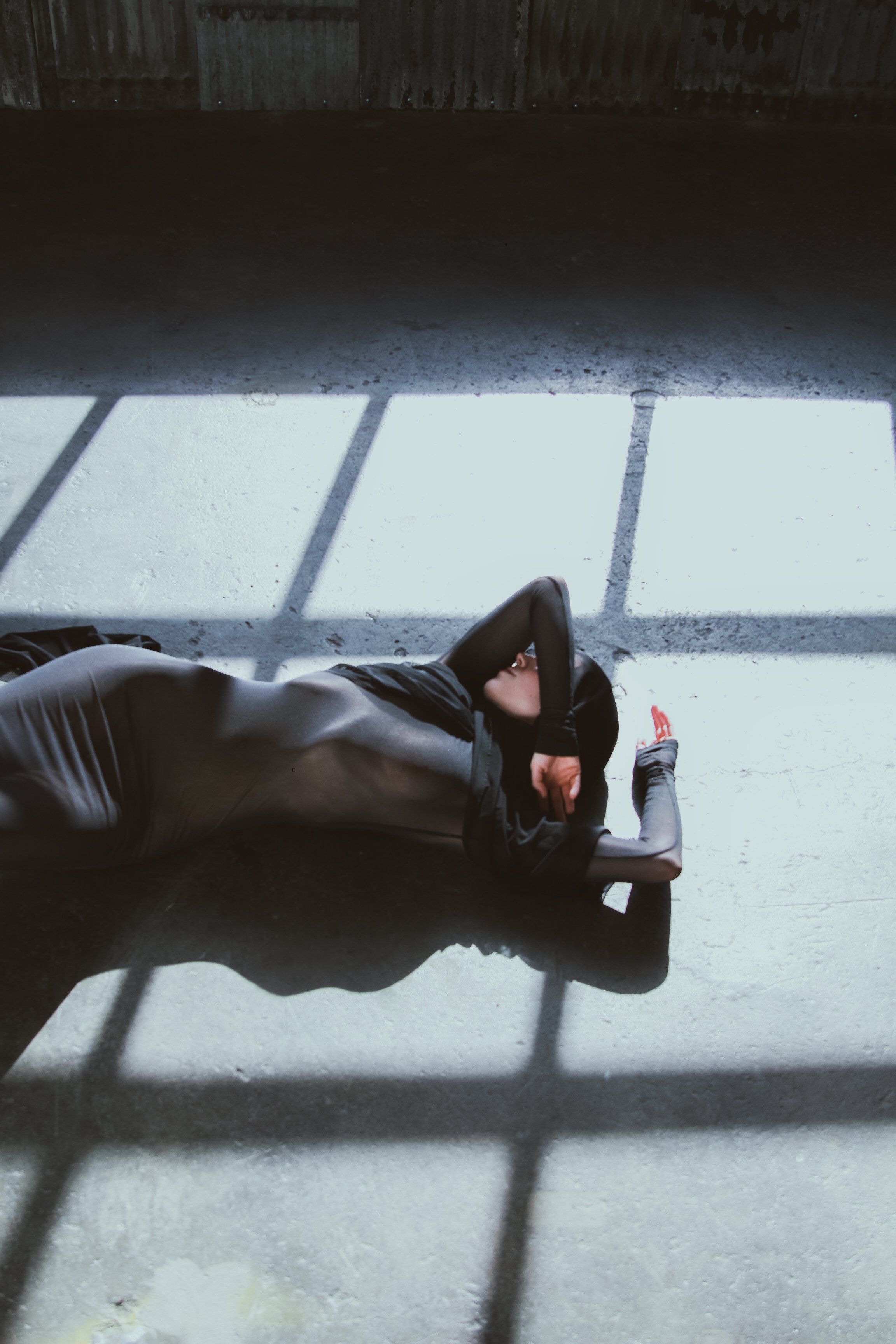 Person in black clothing lying on a concrete floor, with sunlight casting window-shaped shadows.