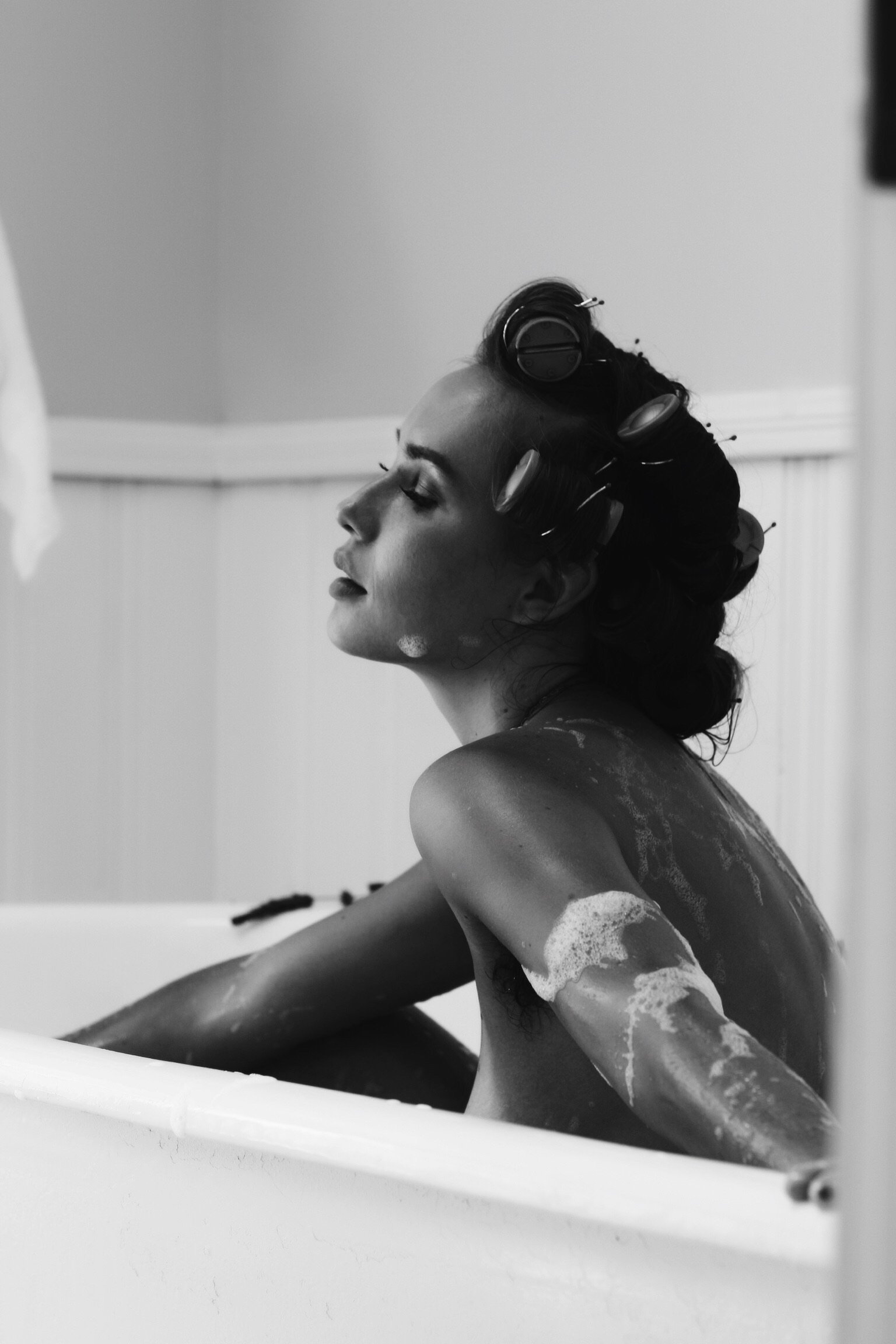 Black and white image of a person with hair curlers, relaxing in a bathtub with soap suds on their skin.