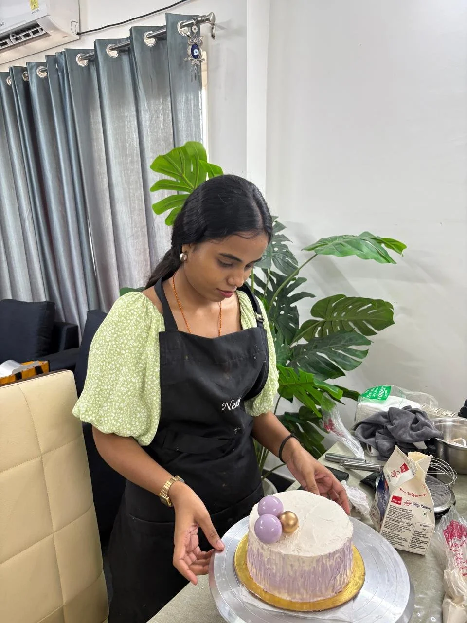 A woman in a black apron and a green dotted blouse stands at a table with a decorated birthday cake. The cake is round with white frosting, a gold base, and three purple and one gold spherical decorations on top. The background includes large green p