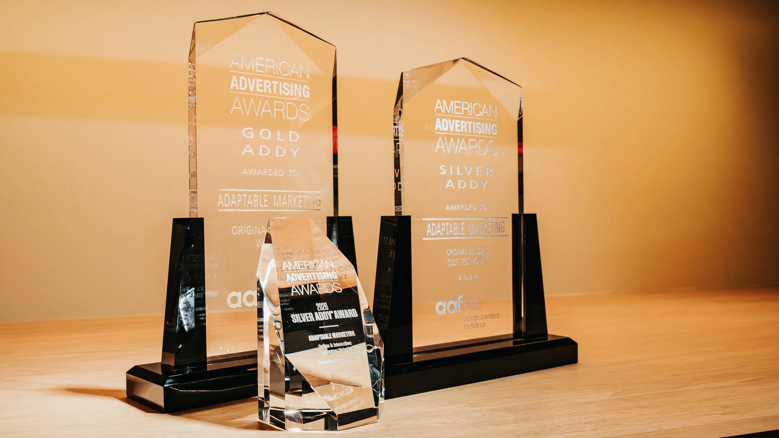 Several glass awards for the American Advertising Awards, including a silver, gold, and smaller plaques, displayed on a wooden surface against a plain wall.