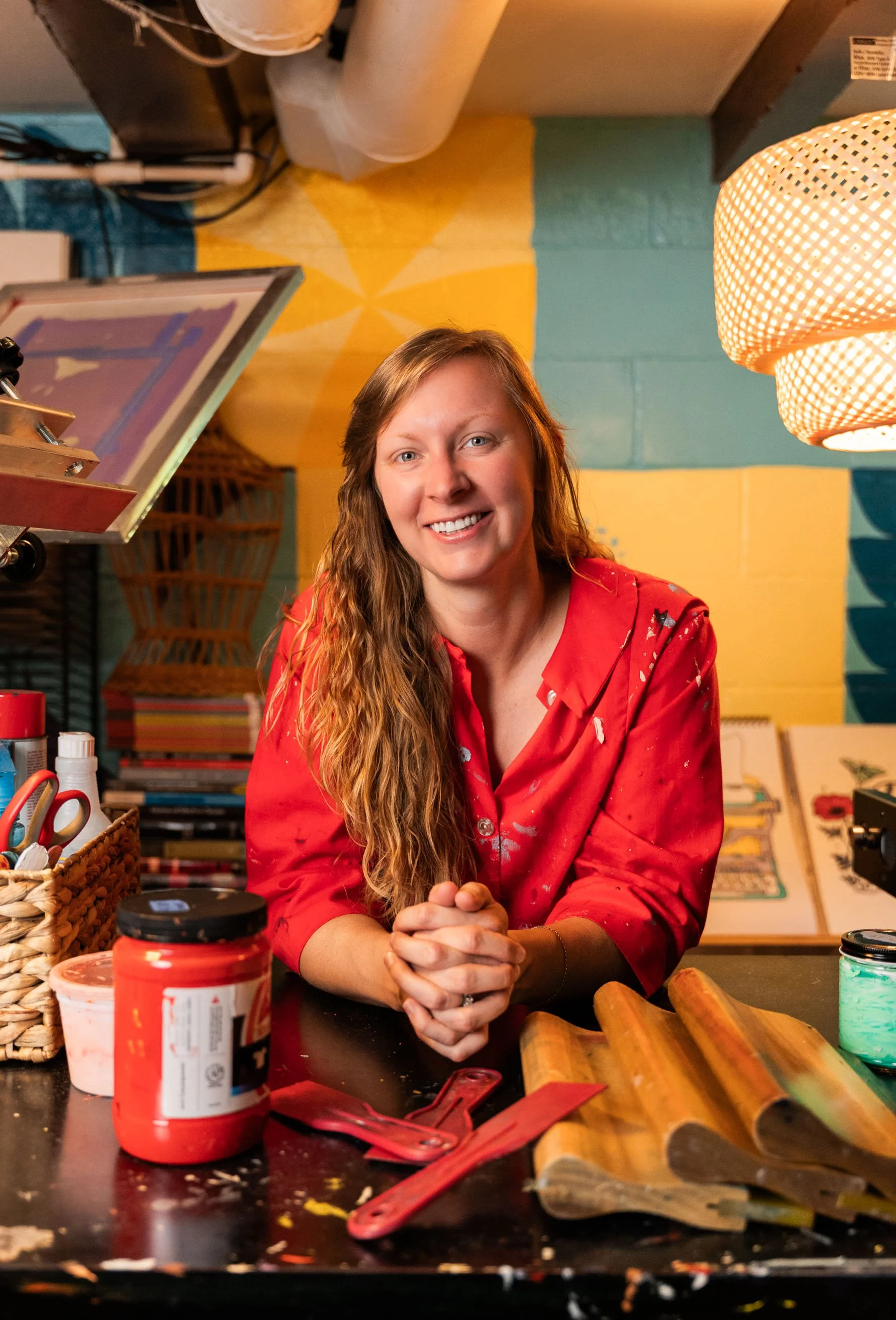 A woman with long curly hair smiling at the camera, sitting at a table in a colorful room with art supplies, a basket, and a light fixture in the background. She is wearing a red shirt and has her hands clasped together.