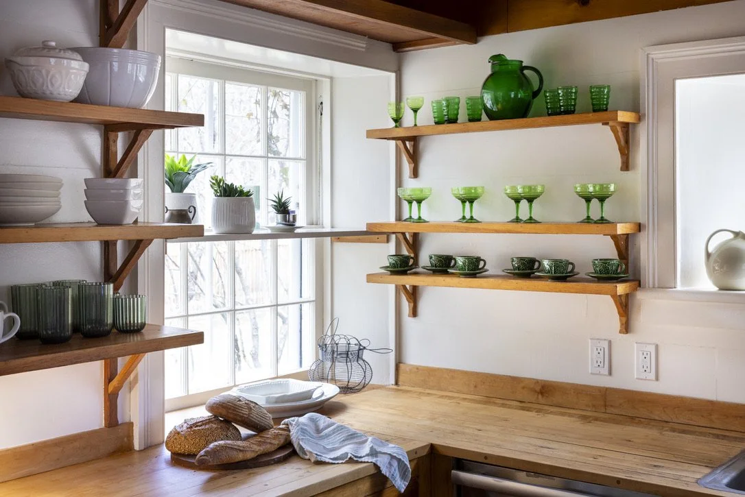 Kitchen staged by Nancy Geaney of NDG Studio with wooden shelves holding white and green dishes, a countertop with bread and a dish towel, and a window with pots and plants outside.