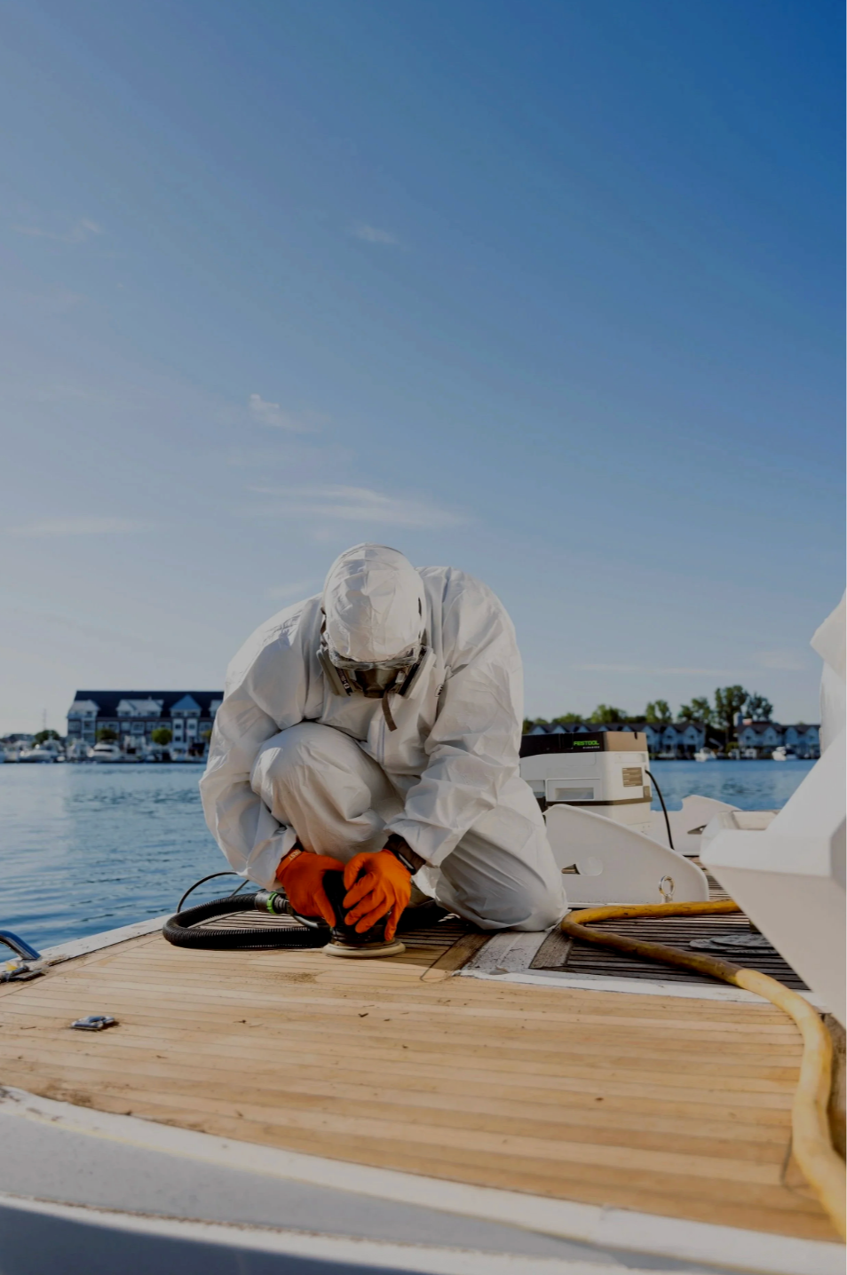 A person wearing protective white coveralls and orange gloves working on a boat deck with equipment near a body of water during daytime.