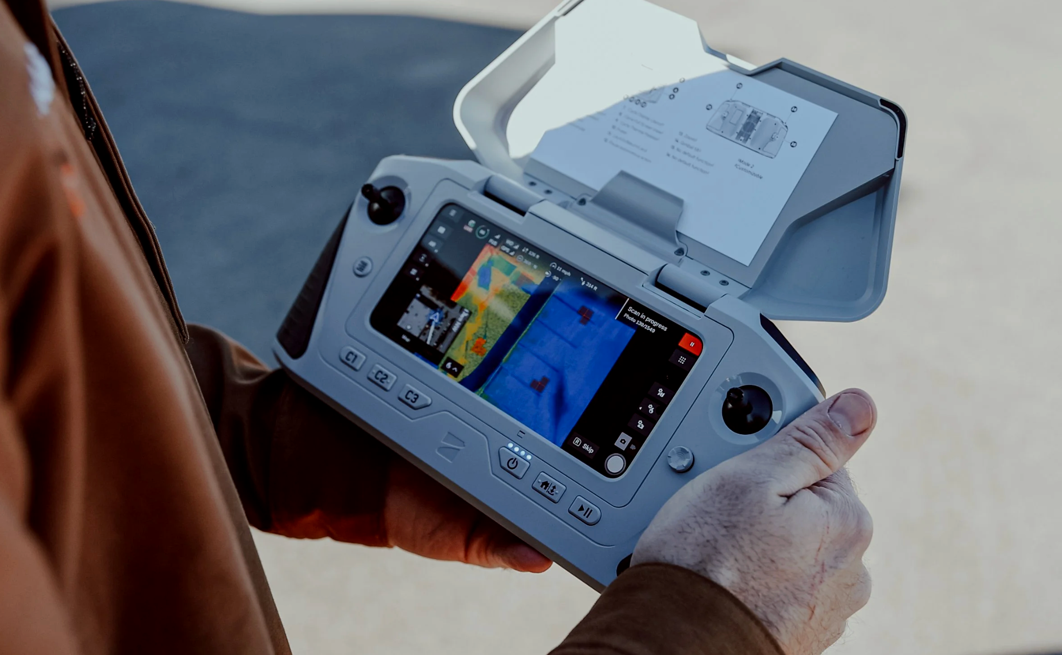 Person holding a drone controller with a screen displaying thermal imaging maps and a printed instruction sheet.