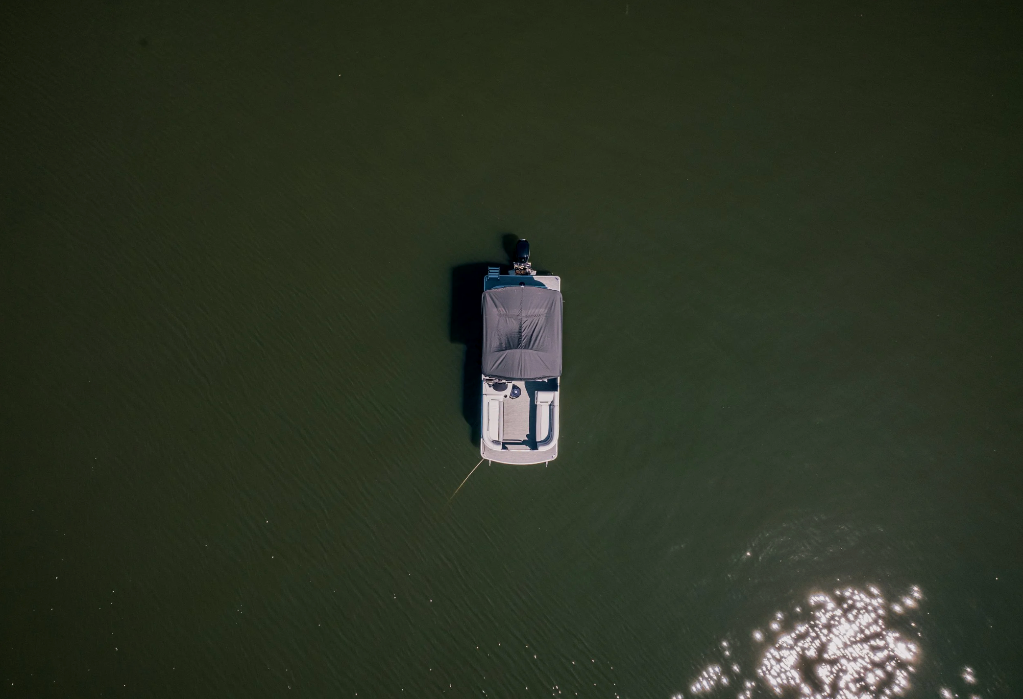 An aerial view of a boat with a black cover on the water, casting a shadow, with sunlight reflections on the water surface.