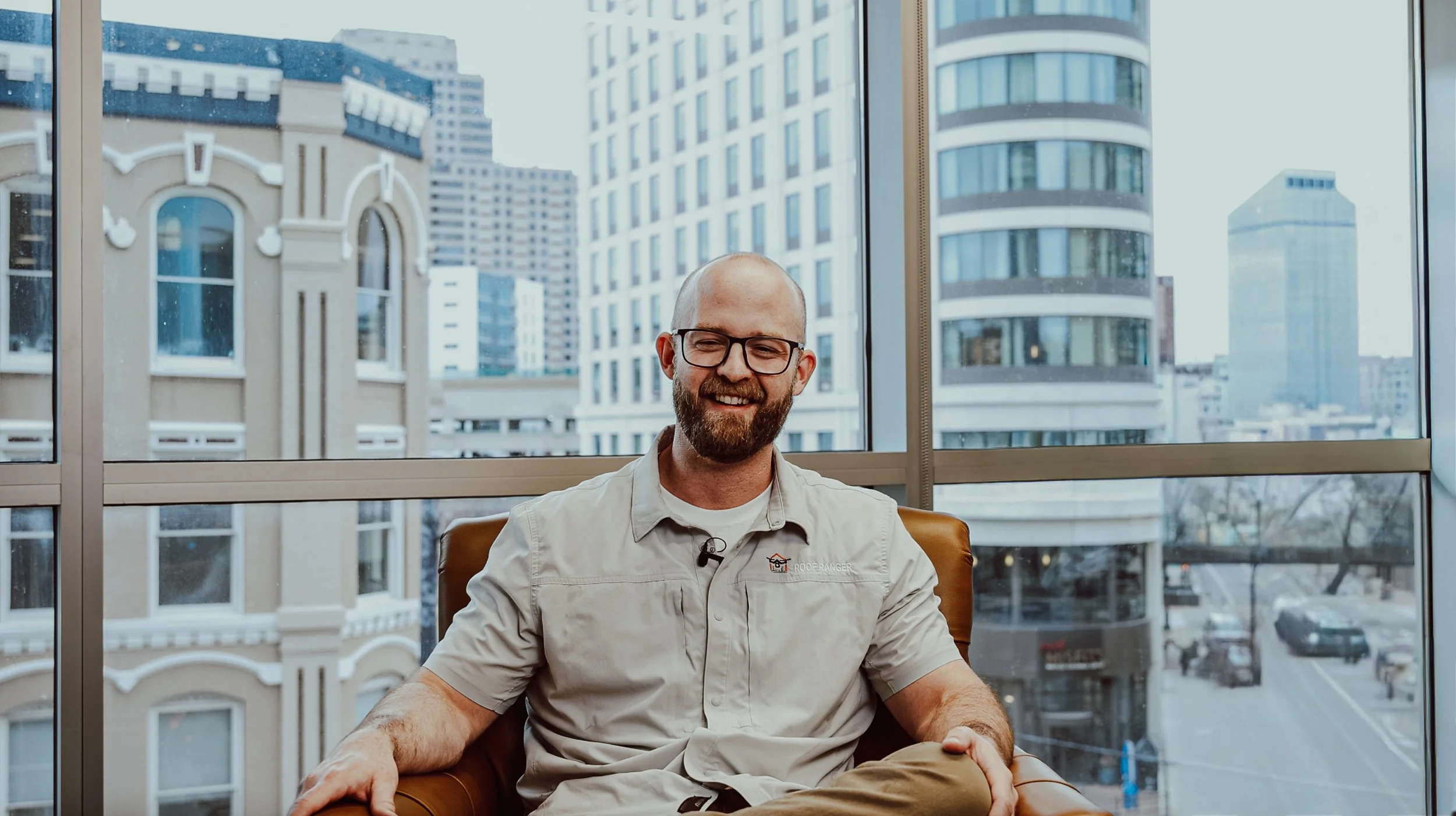 A smiling man with glasses and a beard sitting in a chair in front of large windows with a cityscape background, including tall buildings and streets.