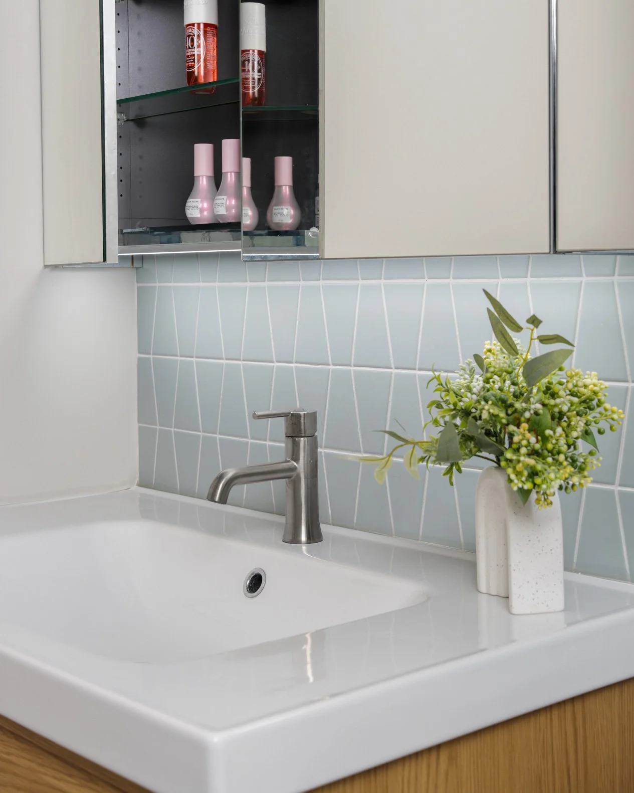 A close-up of a modern bathroom sink area, featuring a white integrated sink and countertop. A brushed nickel faucet is centered over the basin. The backsplash is a light blue diamond-patterned accent tile. Above the sink, a mirrored medicine cabinet