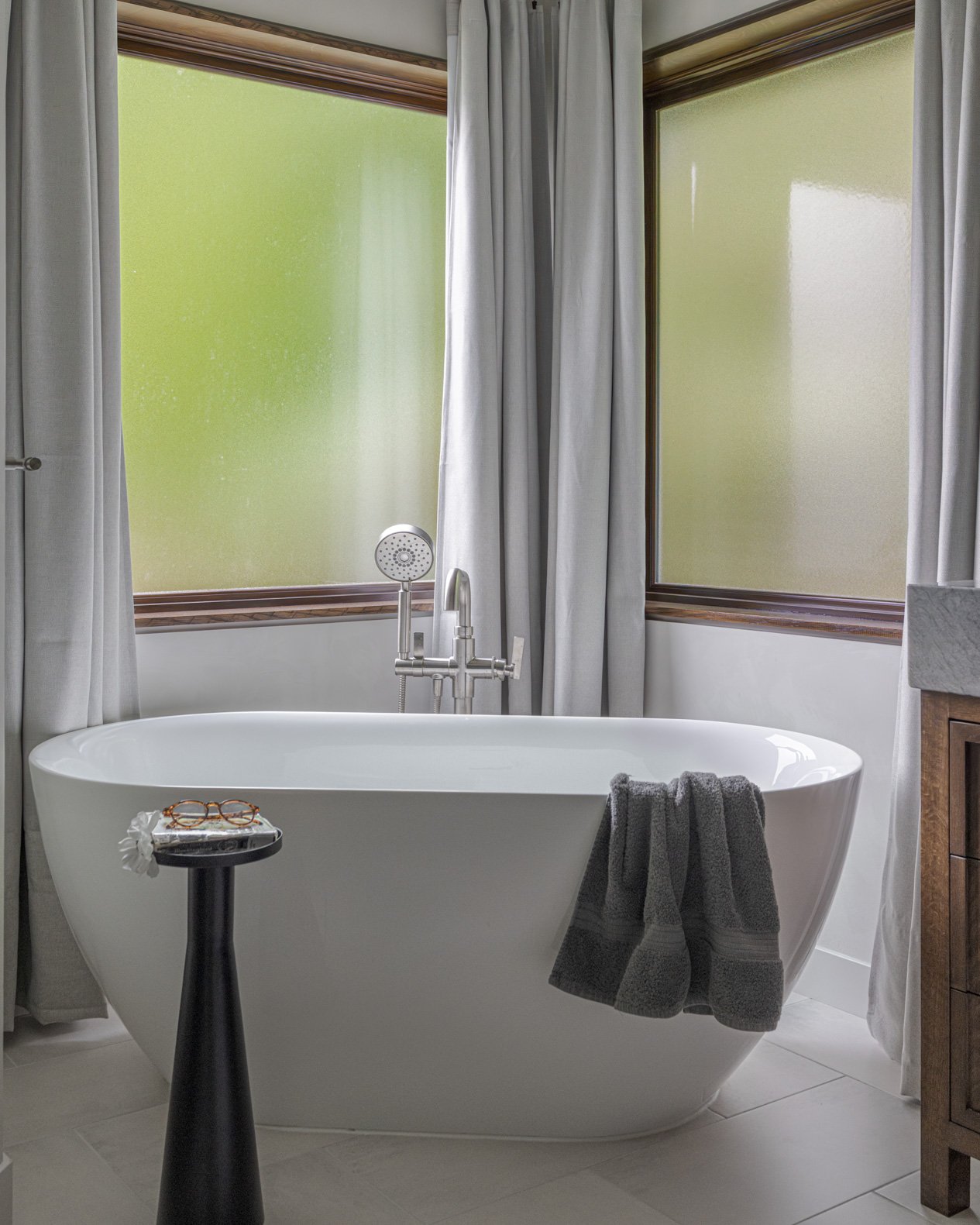 Serene and inviting bathroom featuring a beautiful white freestanding soaking tub positioned elegantly in front of two large windows, offering abundant natural light. The windows are framed with dark wood trim and soft grey curtains, providing privac