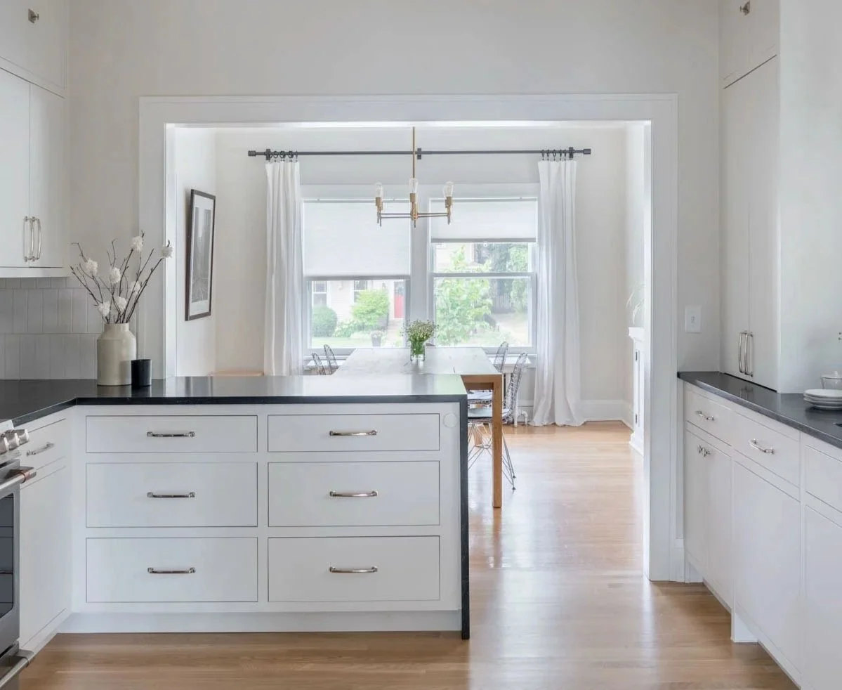 View of the kitchen with white inset cabinets, black countertops, and the architectural cased opening leading into the bright dining room.