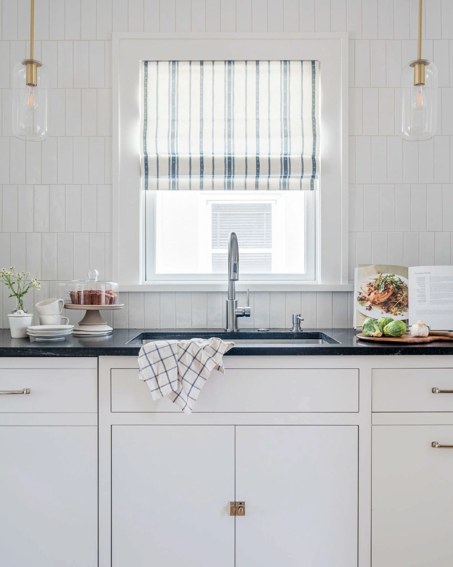 Close-up of kitchen sink area with black countertop, vertical subway tile, white inset cabinetry, and a striped Roman shade.