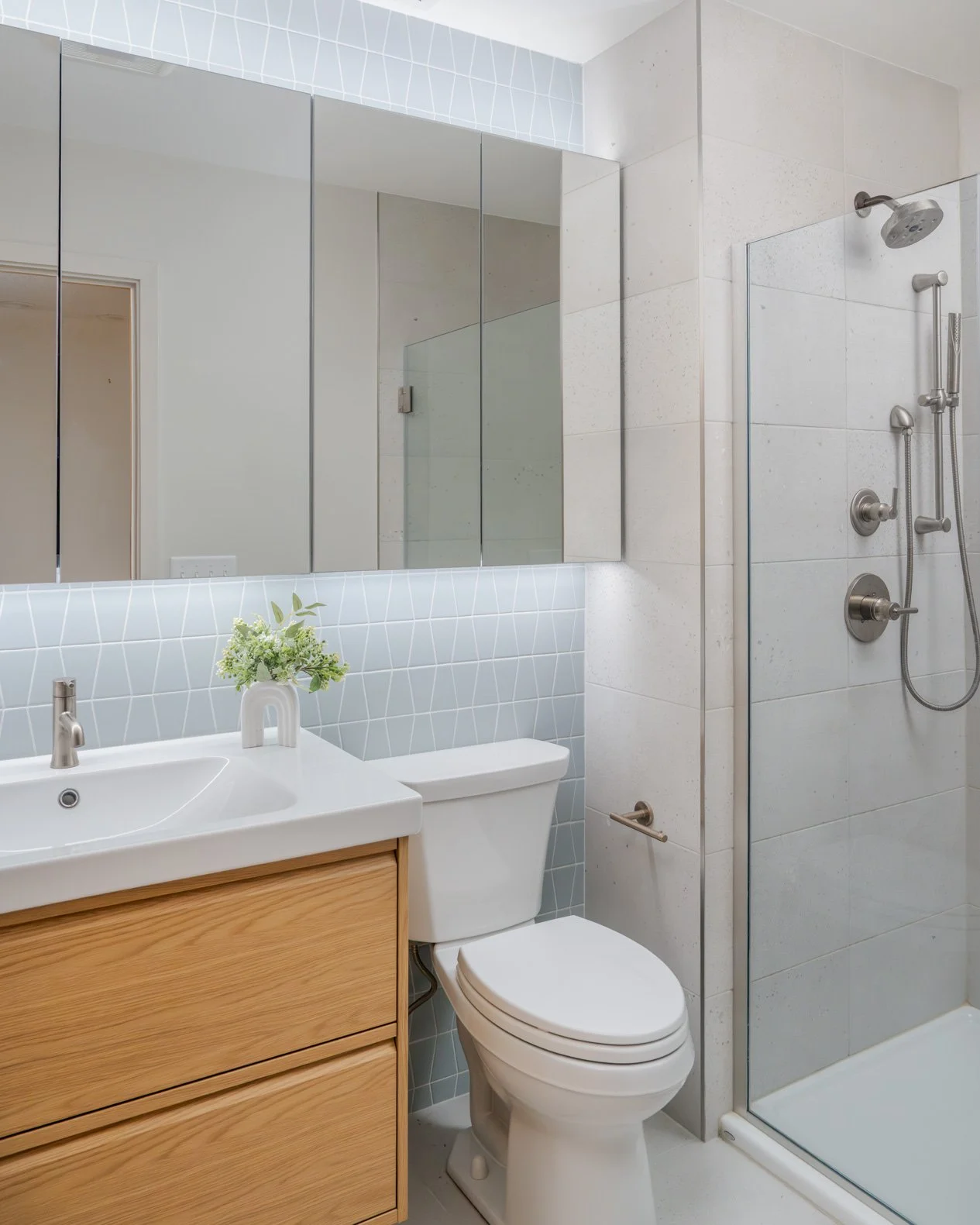 A contemporary bathroom featuring light blue diamond-patterned accent tiles behind a white sink and toilet, a light wood vanity with two drawers, a large mirrored medicine cabinet with integrated lighting, and a walk-in shower with light grey rectang