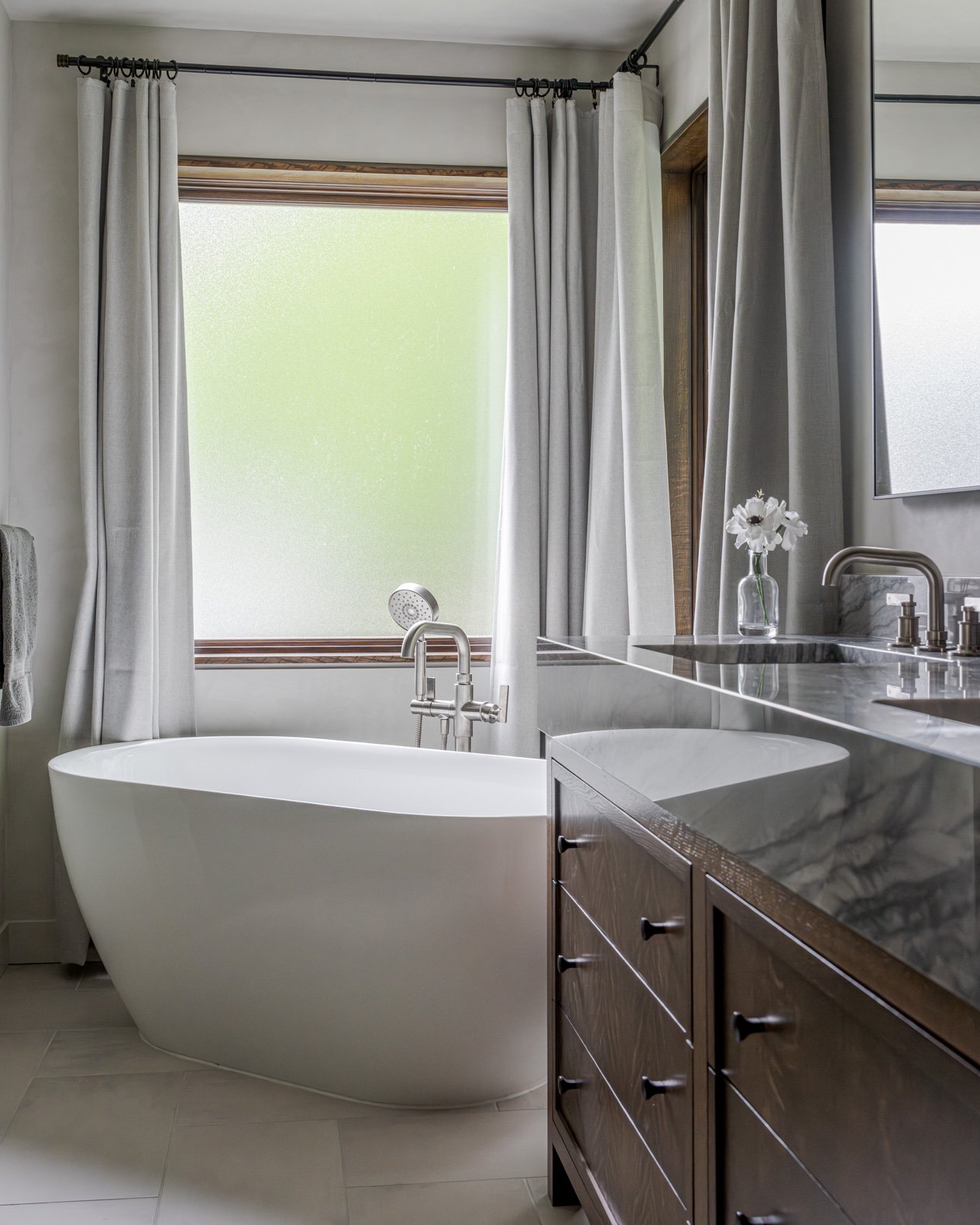 Luxurious freestanding bathtub in front of two windows framed by elegant grey curtains that add texture and soften the space