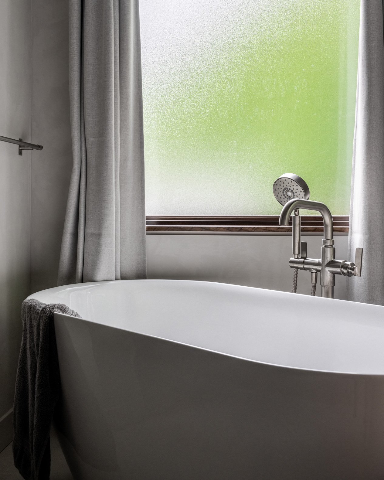 Close-up of a sleek white freestanding bathtub in a modern bathroom, positioned in front of a large frosted window that allows for privacy while bringing in natural light. The tub is equipped with a contemporary brushed nickel floor-mounted tub fille