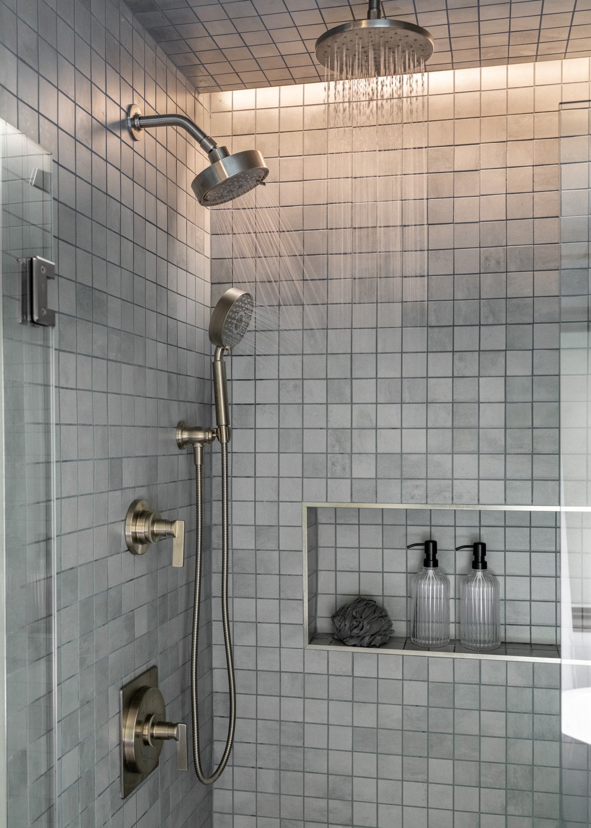 Modern and sleek walk-in shower featuring light grey square tiles throughout, creating a clean and contemporary look. The shower is equipped with both a ceiling-mounted rainfall showerhead and a wall-mounted handheld shower, both in a brushed nickel
