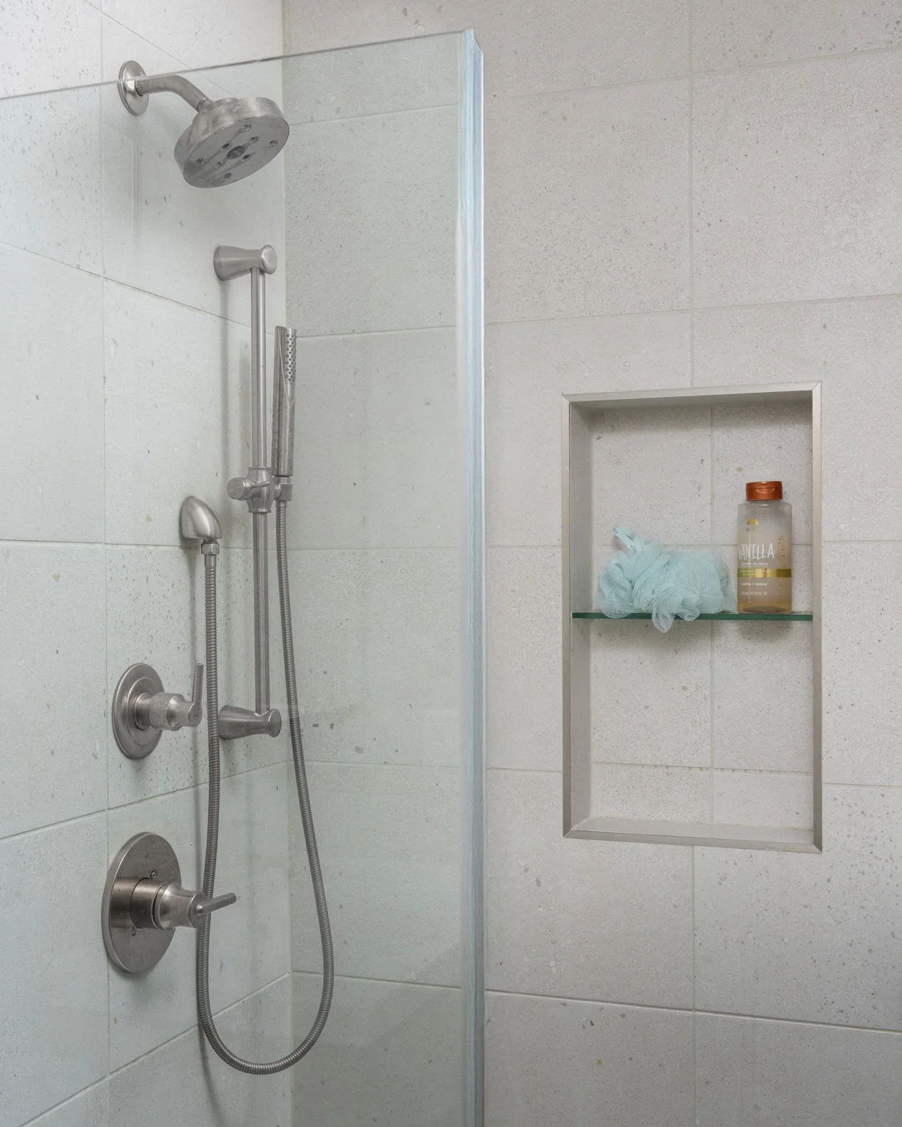 A contemporary shower stall with large, light grey speckled rectangular tiles. The shower features a brushed nickel shower head and a handheld sprayer on an adjustable bar. A vertical recessed niche with a glass shelf is built into the tile wall, hol