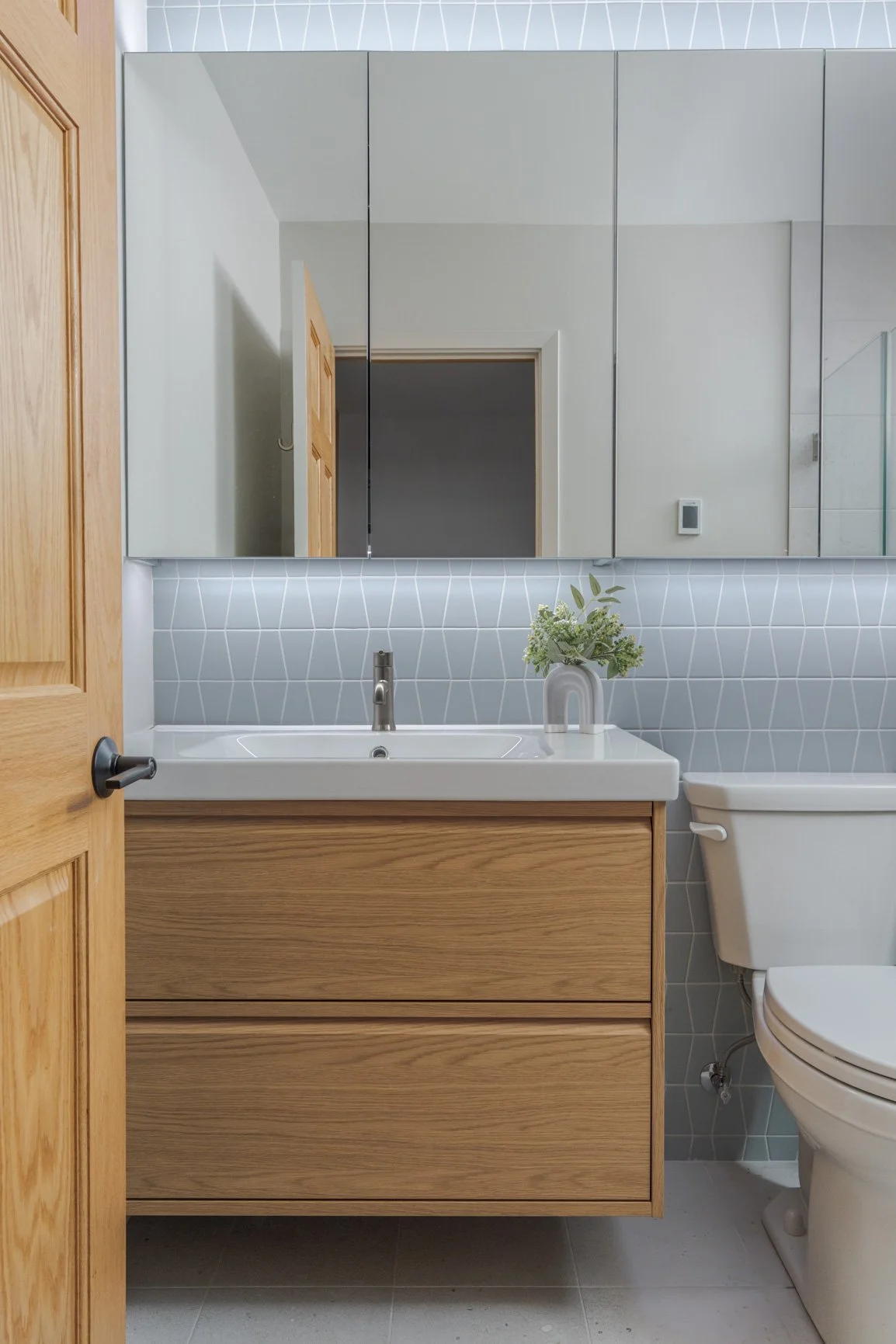 A contemporary bathroom featuring a white oak floating vanity with two drawers, a white sink, and a light blue tapezoid-patterned accent tile backsplash. A large mirrored medicine cabinet with under-cabinet lighting hangs above the sink. The toilet i