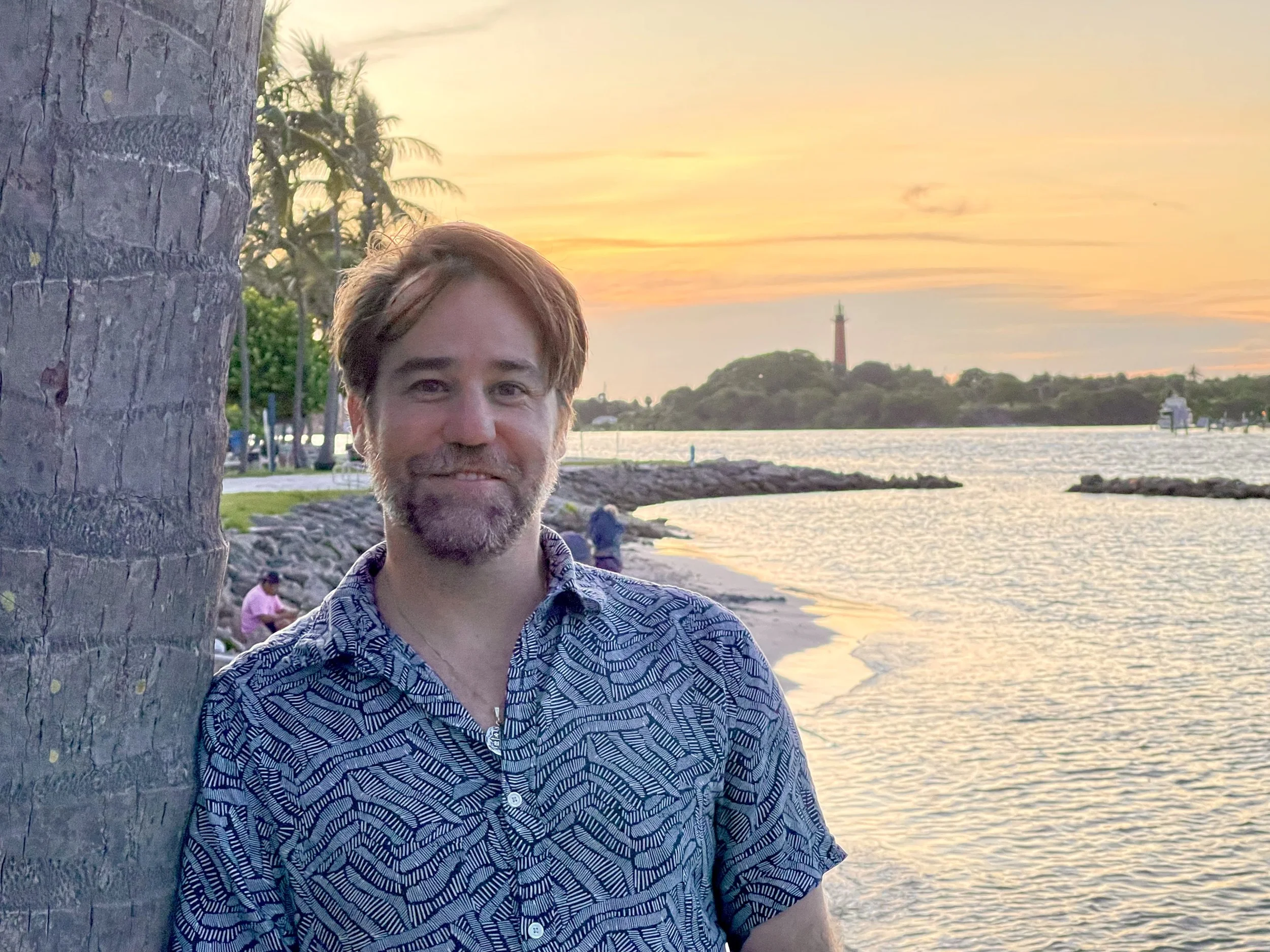 Person standing by a palm tree near a waterfront during sunset; lighthouse and distant trees in background.