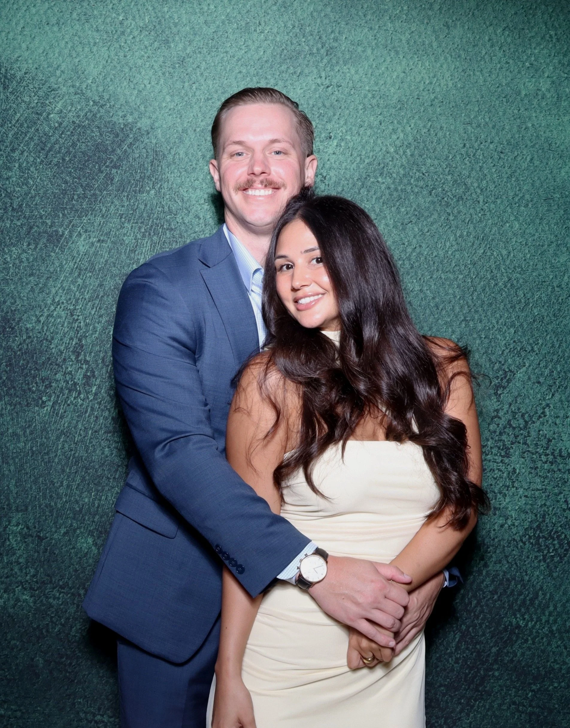 A smiling couple, a man in a blue suit and a woman in a white dress, embracing against a textured green background.