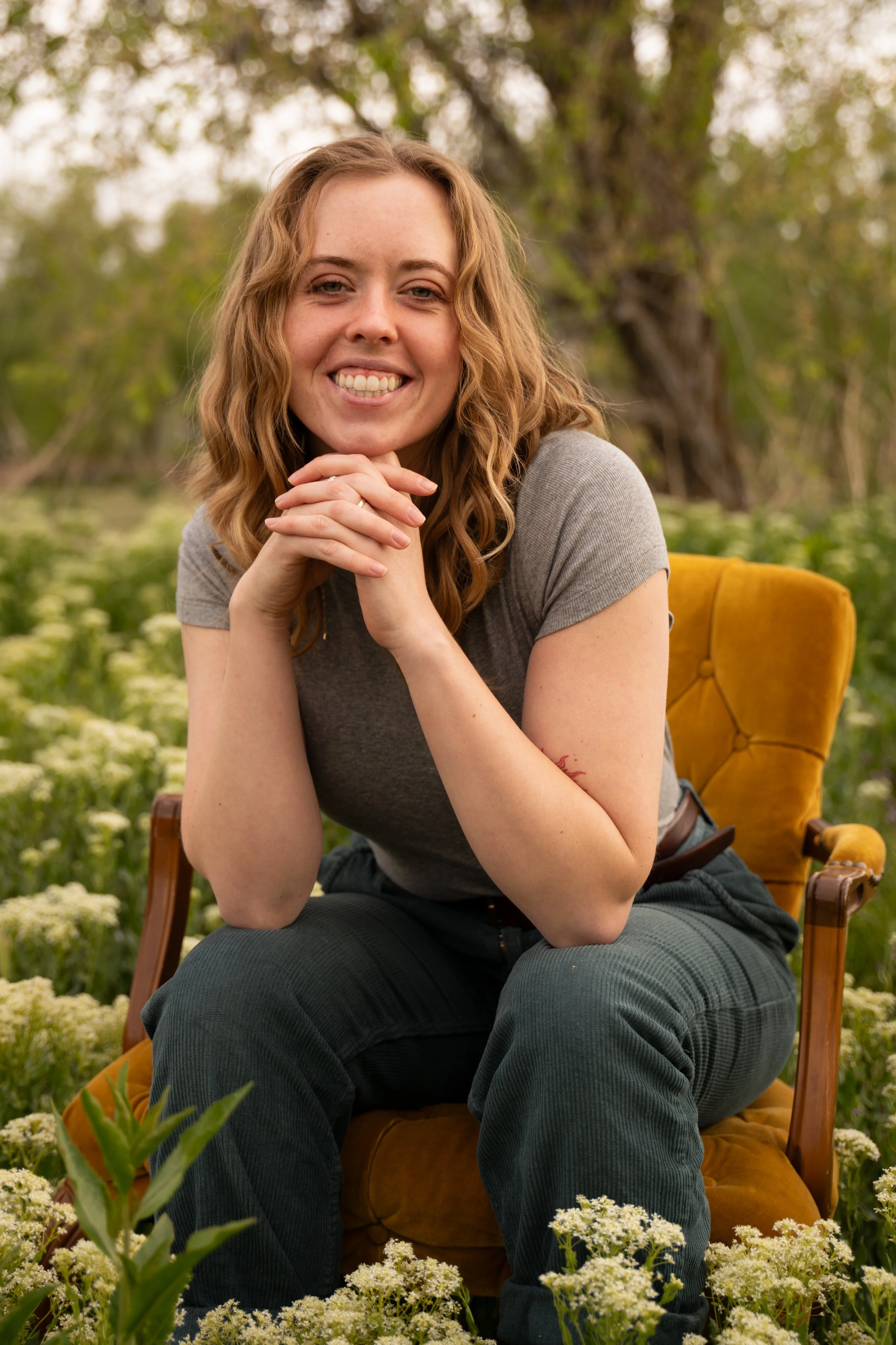 A woman sitting on a yellow chair outdoors surrounded by white flowers and trees, smiling with her hands clasped under her chin.