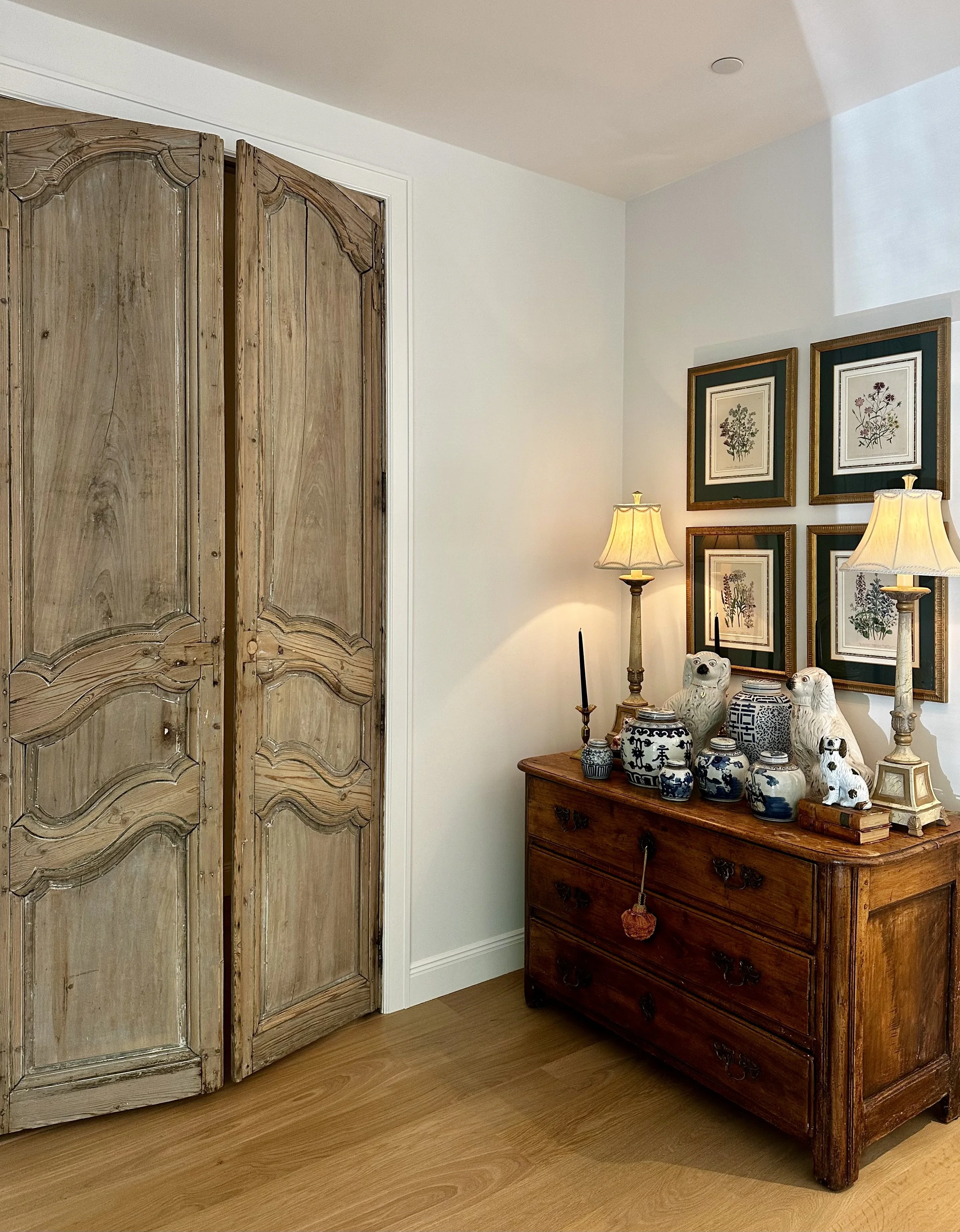 Antique wood doors beside a collected vignette of botanical artwork, blue-and-white pottery, lamps, and an antique chest.