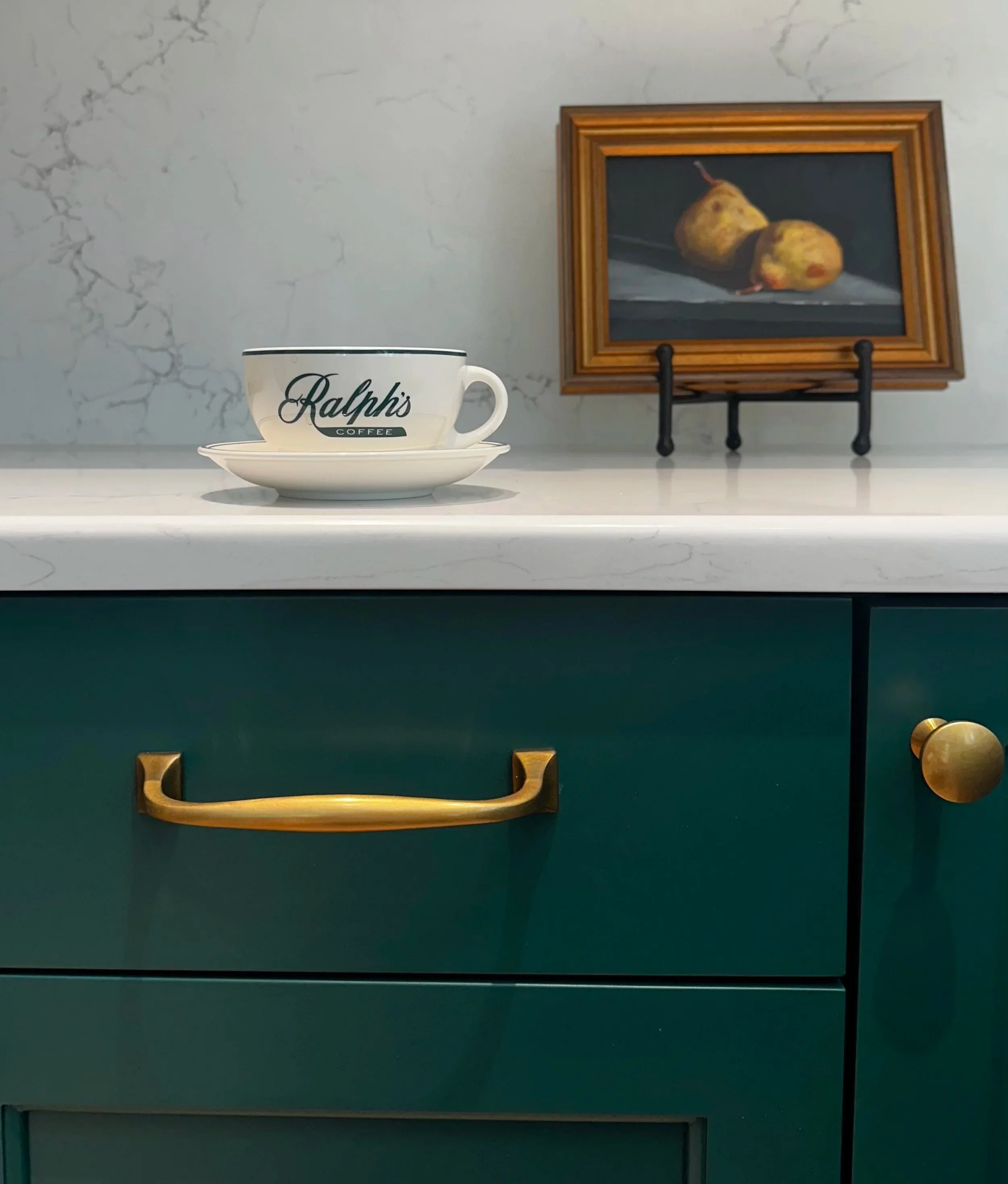 Ralph’s Coffee cup displayed above deep green kitchen cabinetry with aged brass hardware and a small framed still-life painting.