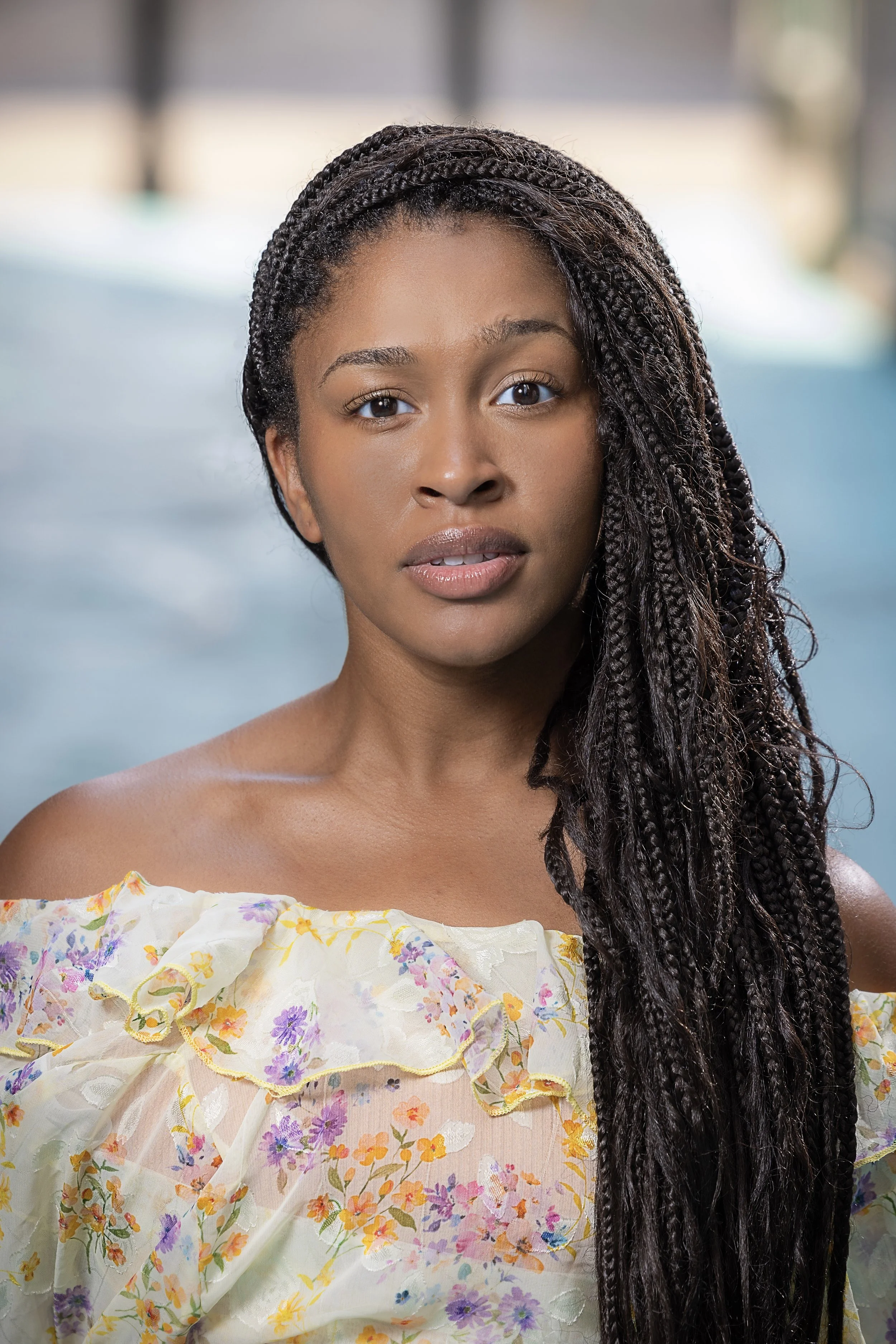 A young woman with long dark braids wearing an off-shoulder floral top, standing confidently outdoors.