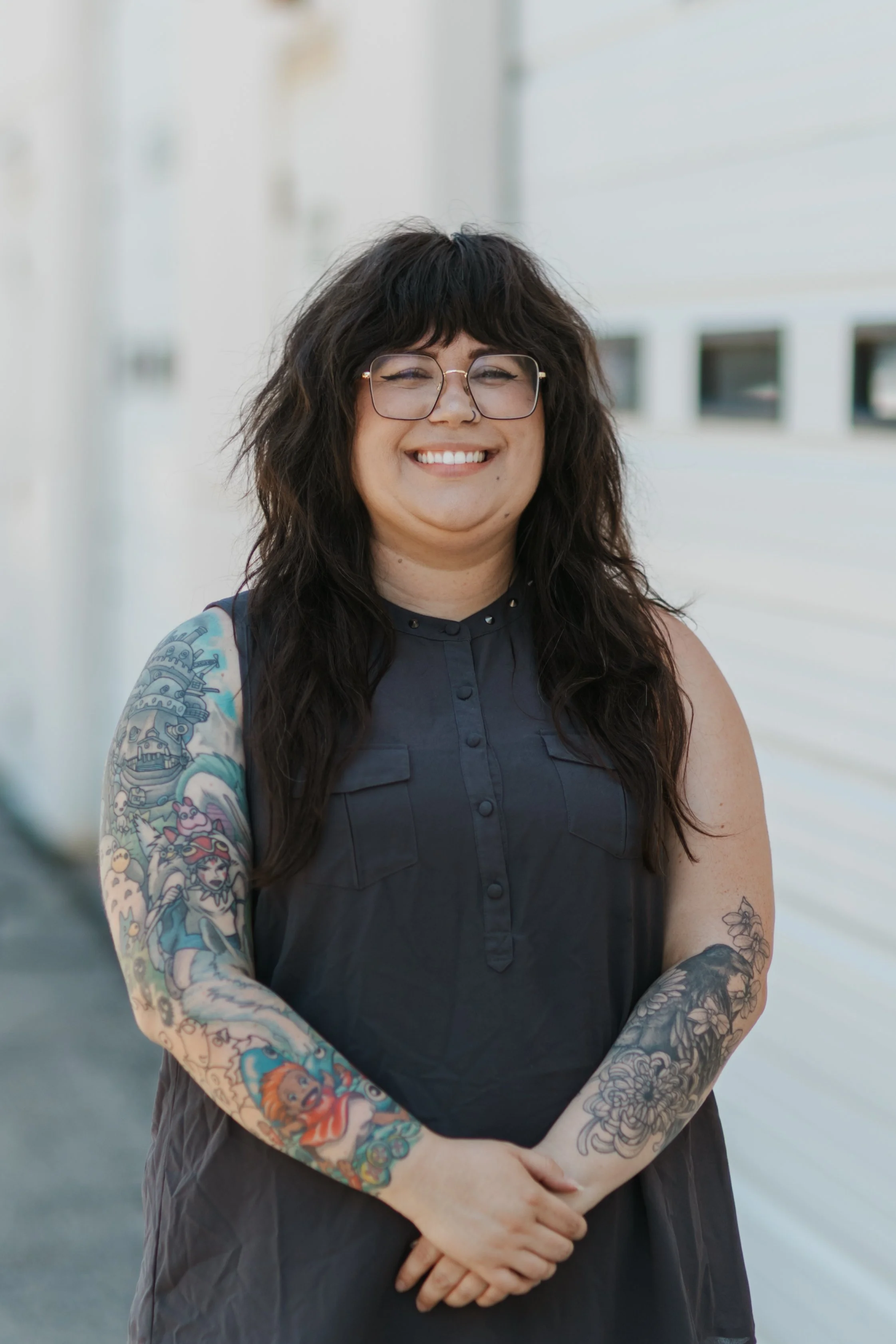 A woman with wavy dark hair, glasses, and a tattoo on her left arm, smiling outdoors in front of a staircase and greenery.