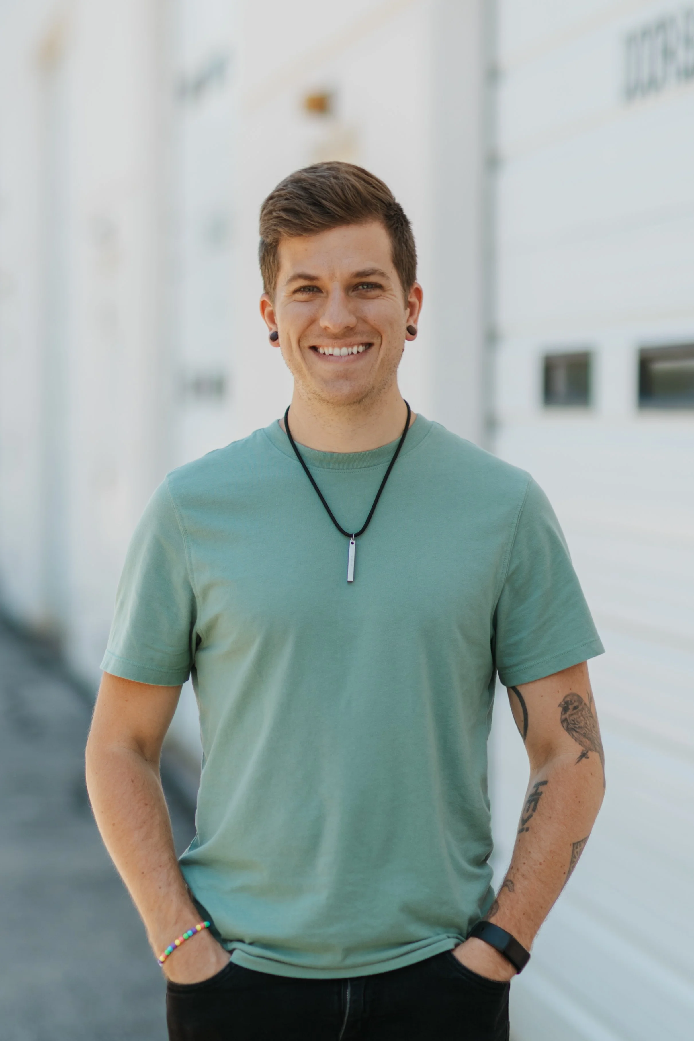 A smiling young man with short brown hair, glasses, and a black jacket over a blue checkered shirt outdoors.