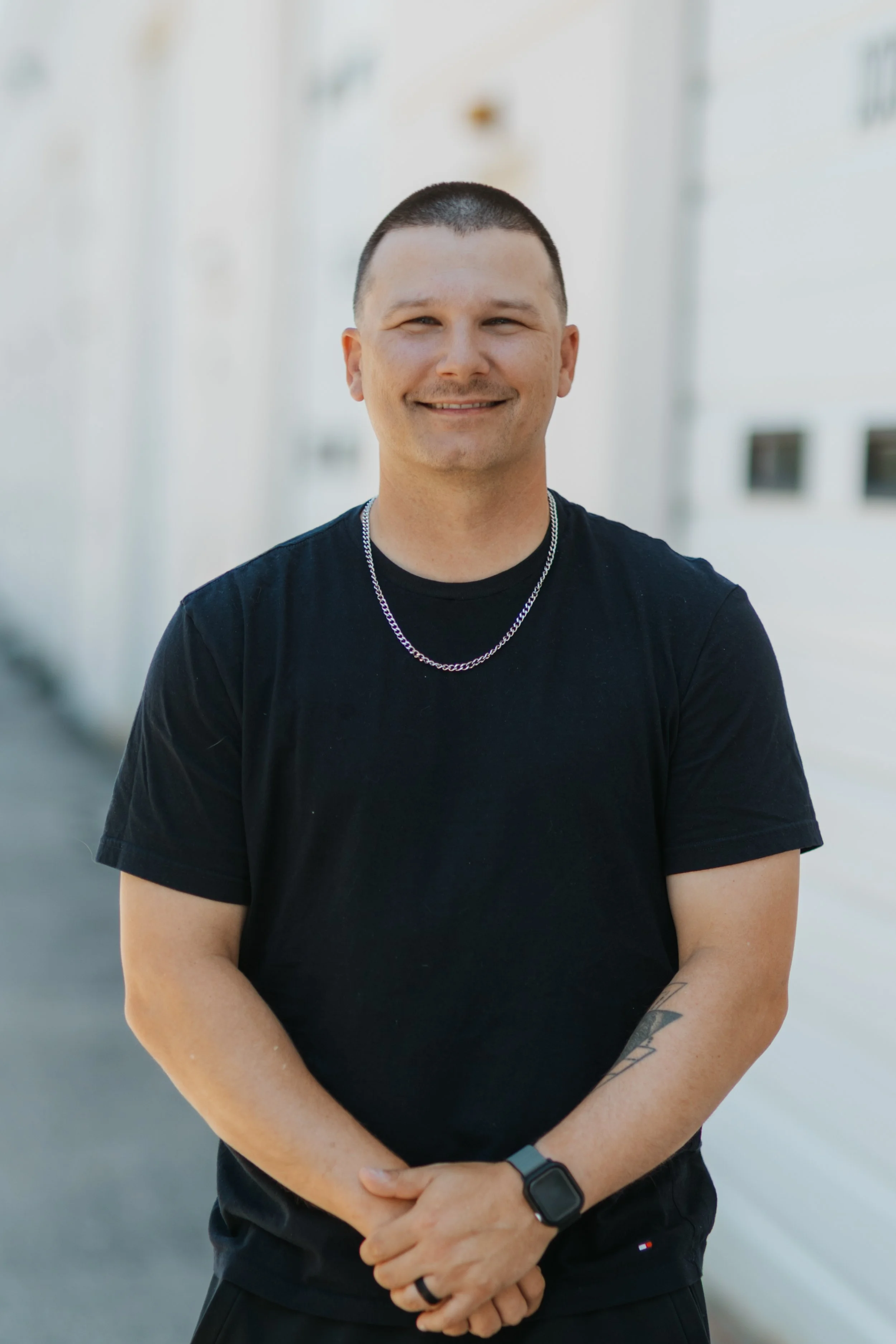 A man smiling outdoors, wearing a blue polo shirt with light blue accents, a white wristwatch, and a tattoo on his right forearm, standing in front of a building and stairs with greenery.