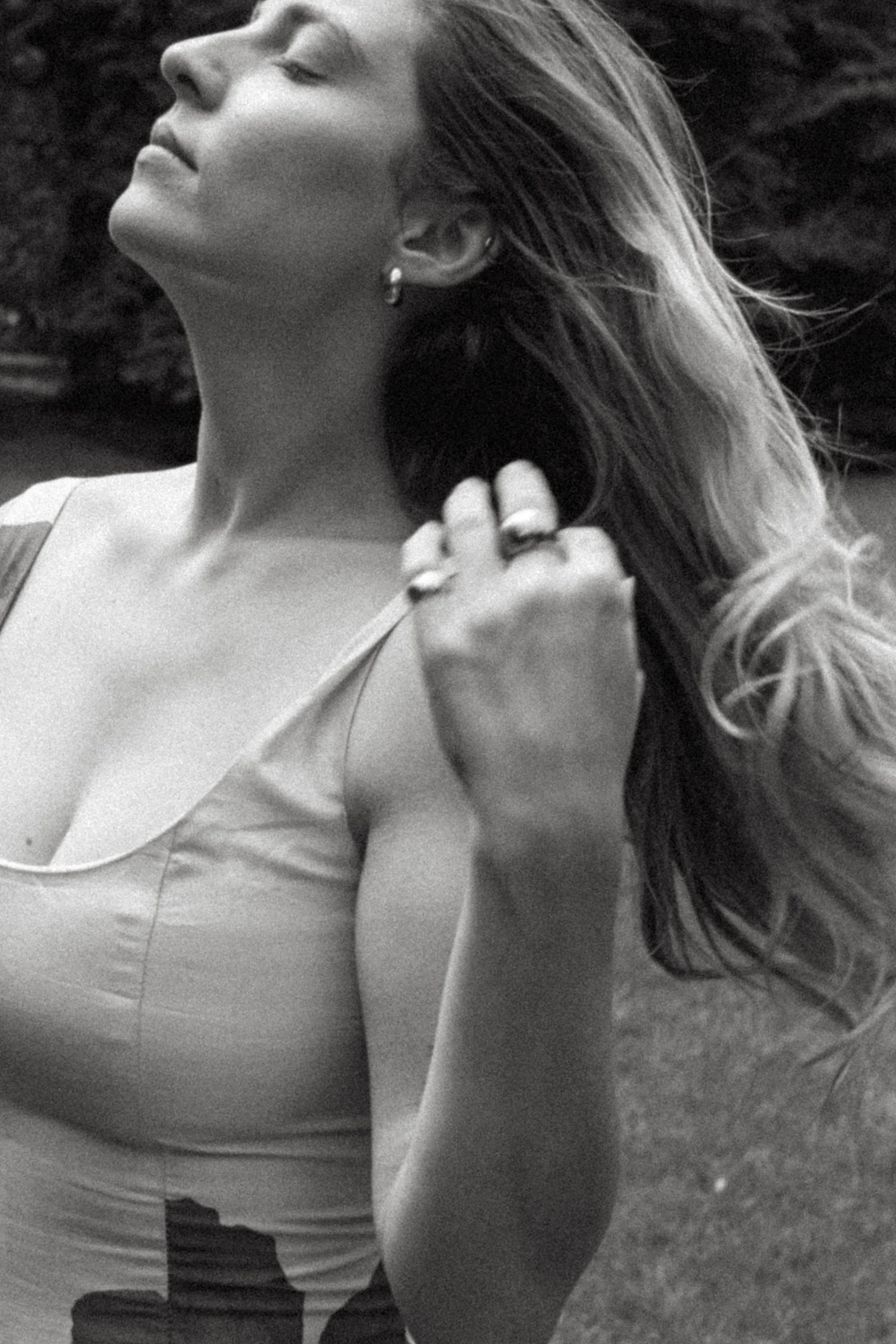 A female wedding photographer with long hair, wearing a sleeveless top, is outdoors with trees in the background. She is touching her hair with her left hand.