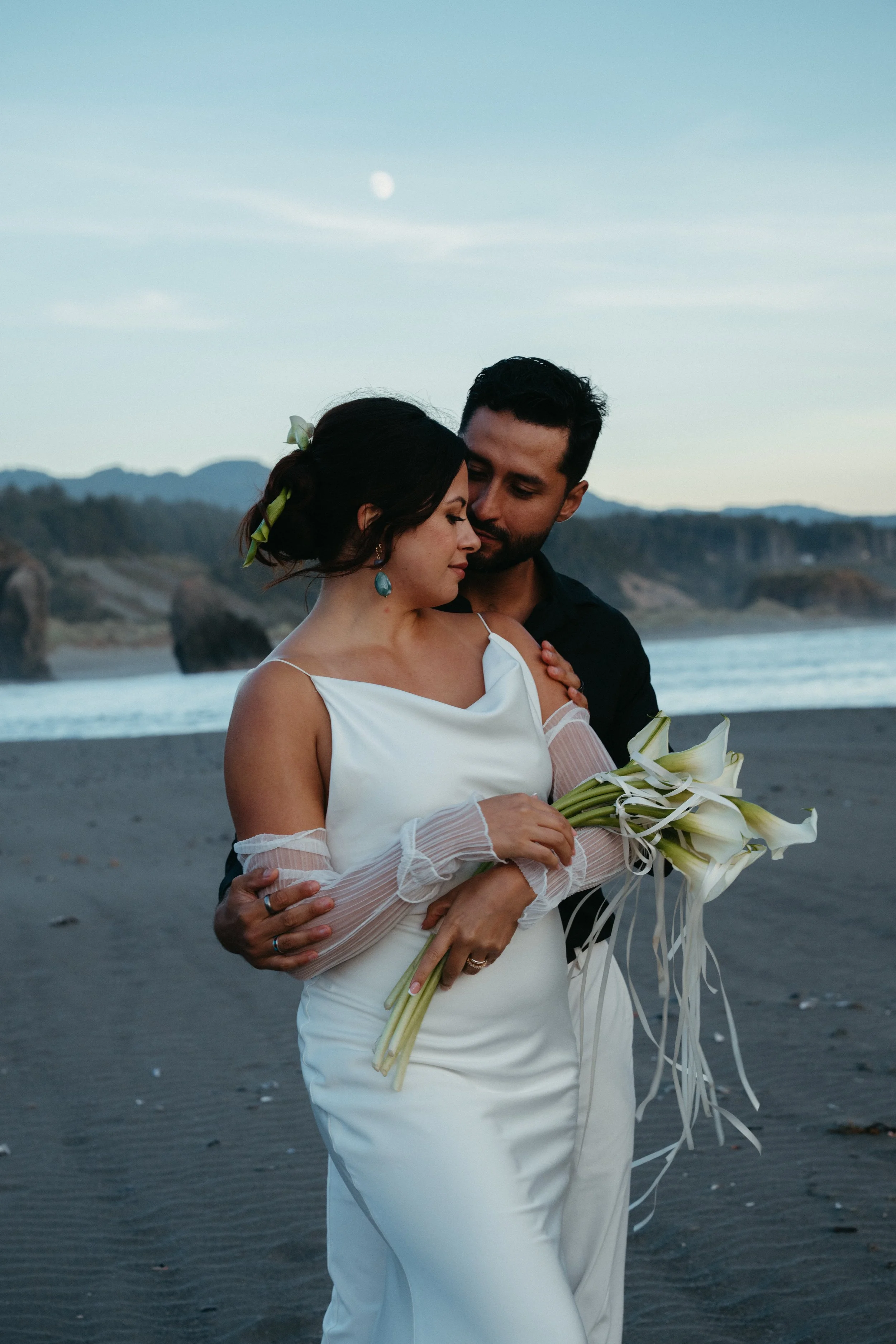 A bride and groom dressed in wedding attire embraces on a beach at sunset, with mountains and the moon in the background, and the woman holding a bouquet of white calla lilies.