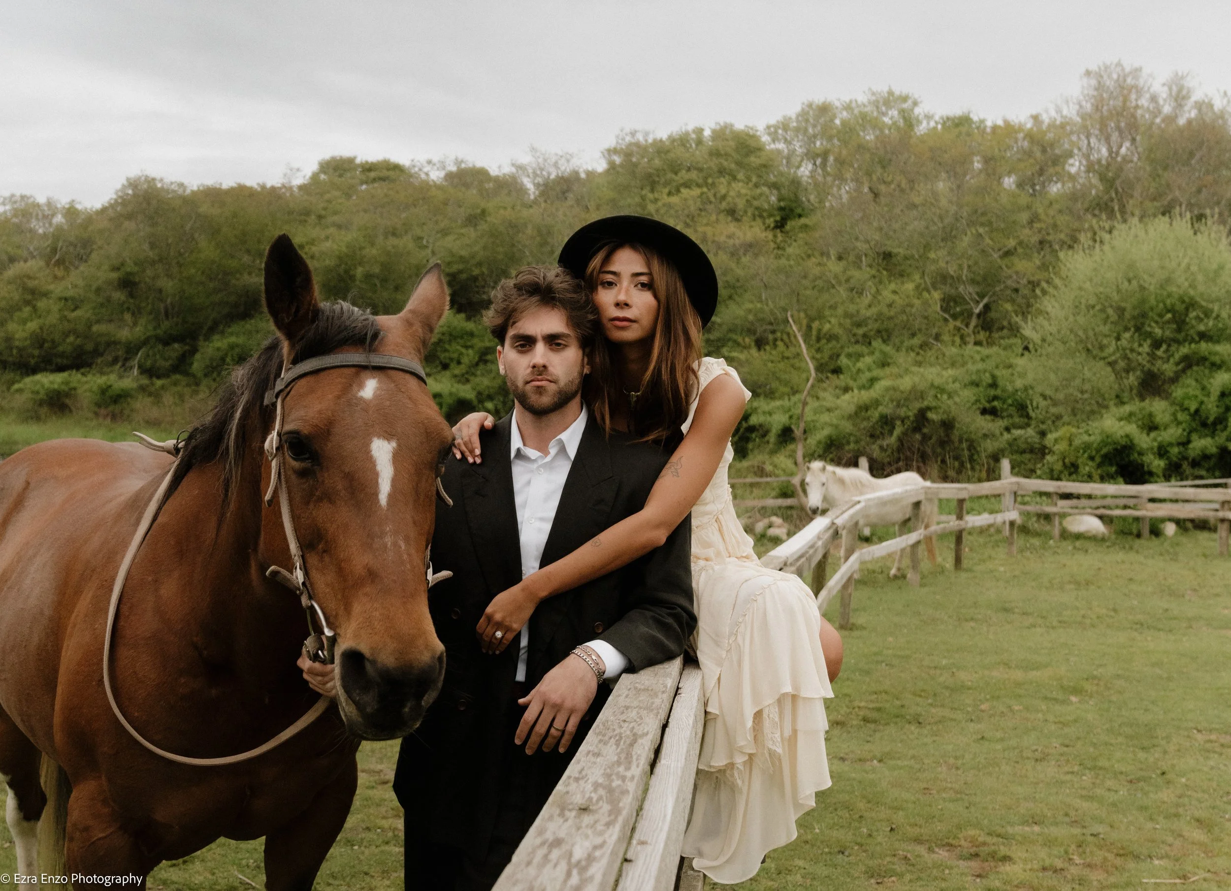 bride sits on top of a wooden fence with her arms around the shoulders of her husband, the groom, and he holds the reins to a brown horse. The couple is within a green meadow and look straight at the photographer