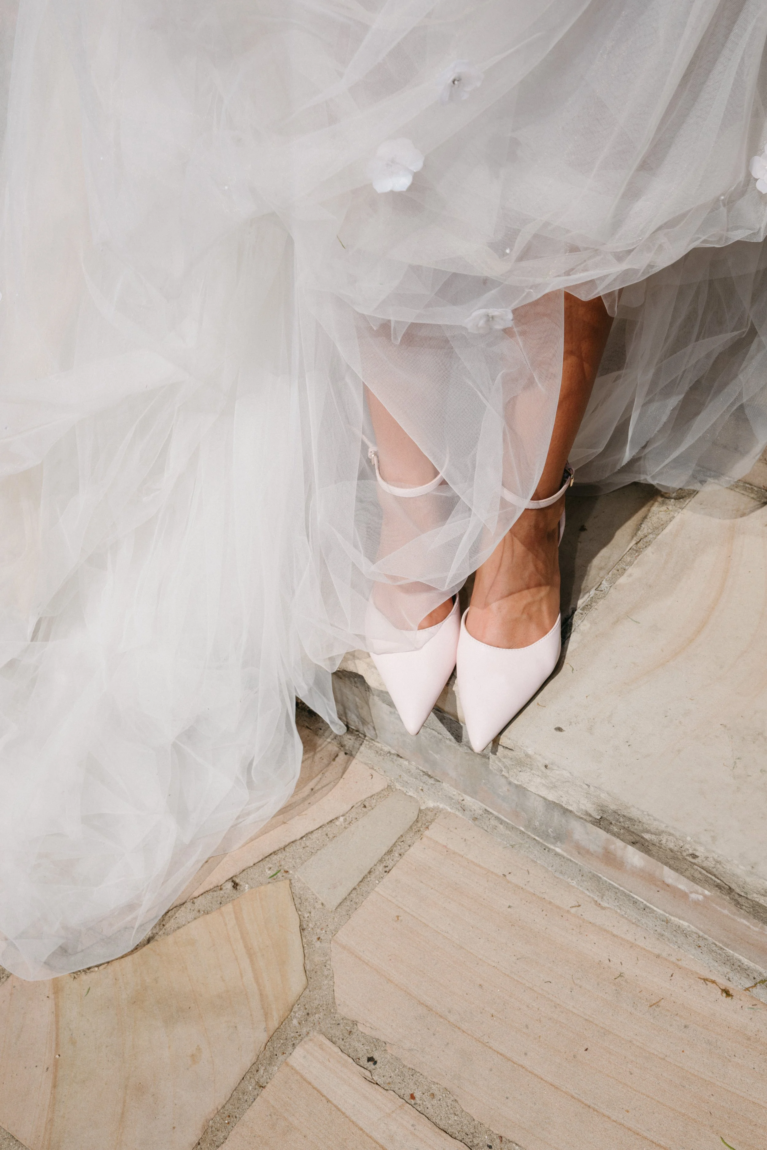 Close-up of a bride's white high heels and garter underneath her layered white tulle wedding dress, standing on a stone-tiled floor.