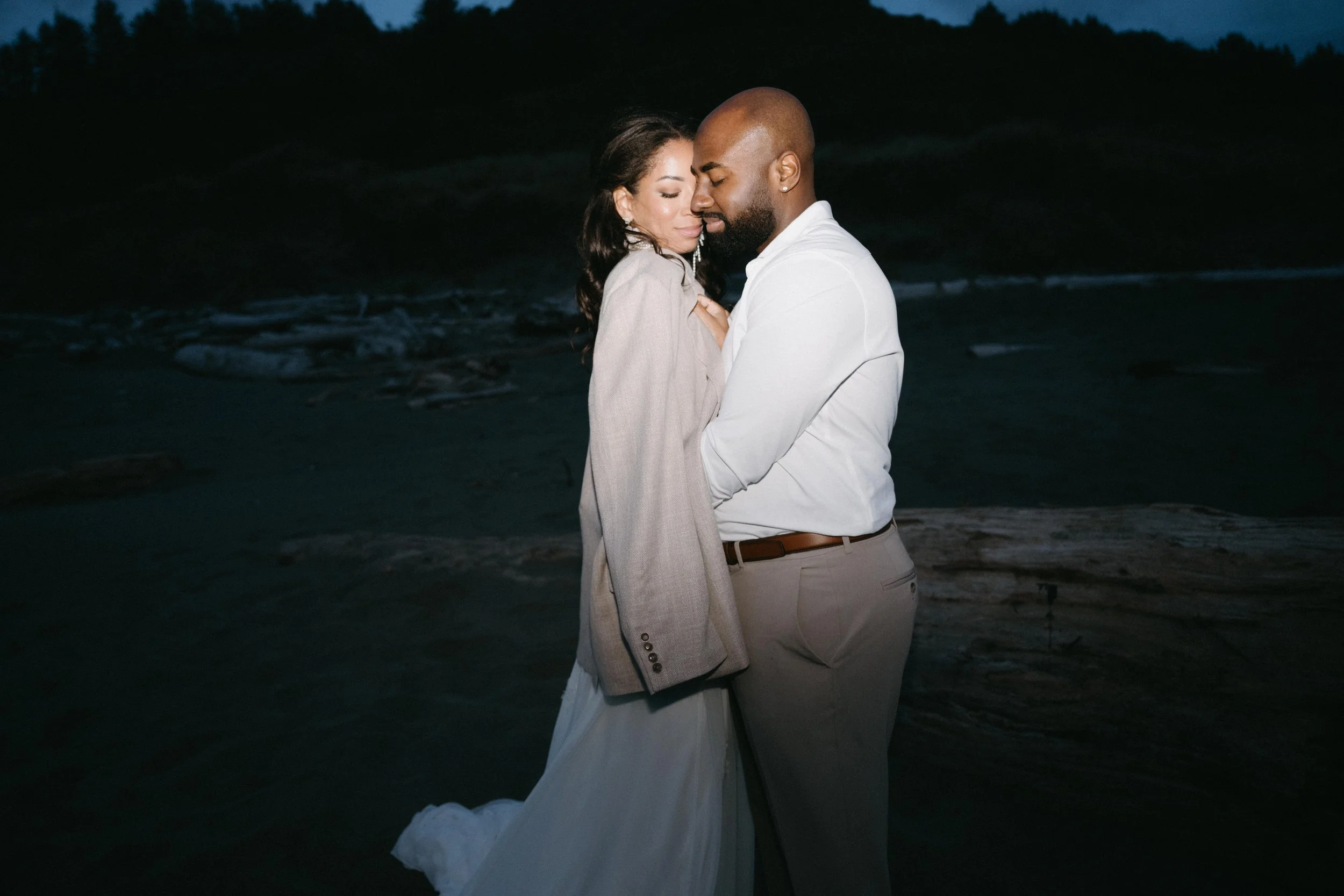 A couple embraces on a beach at dusk at their elopement, with their foreheads touching and eyes closed, dressed in wedding attire.