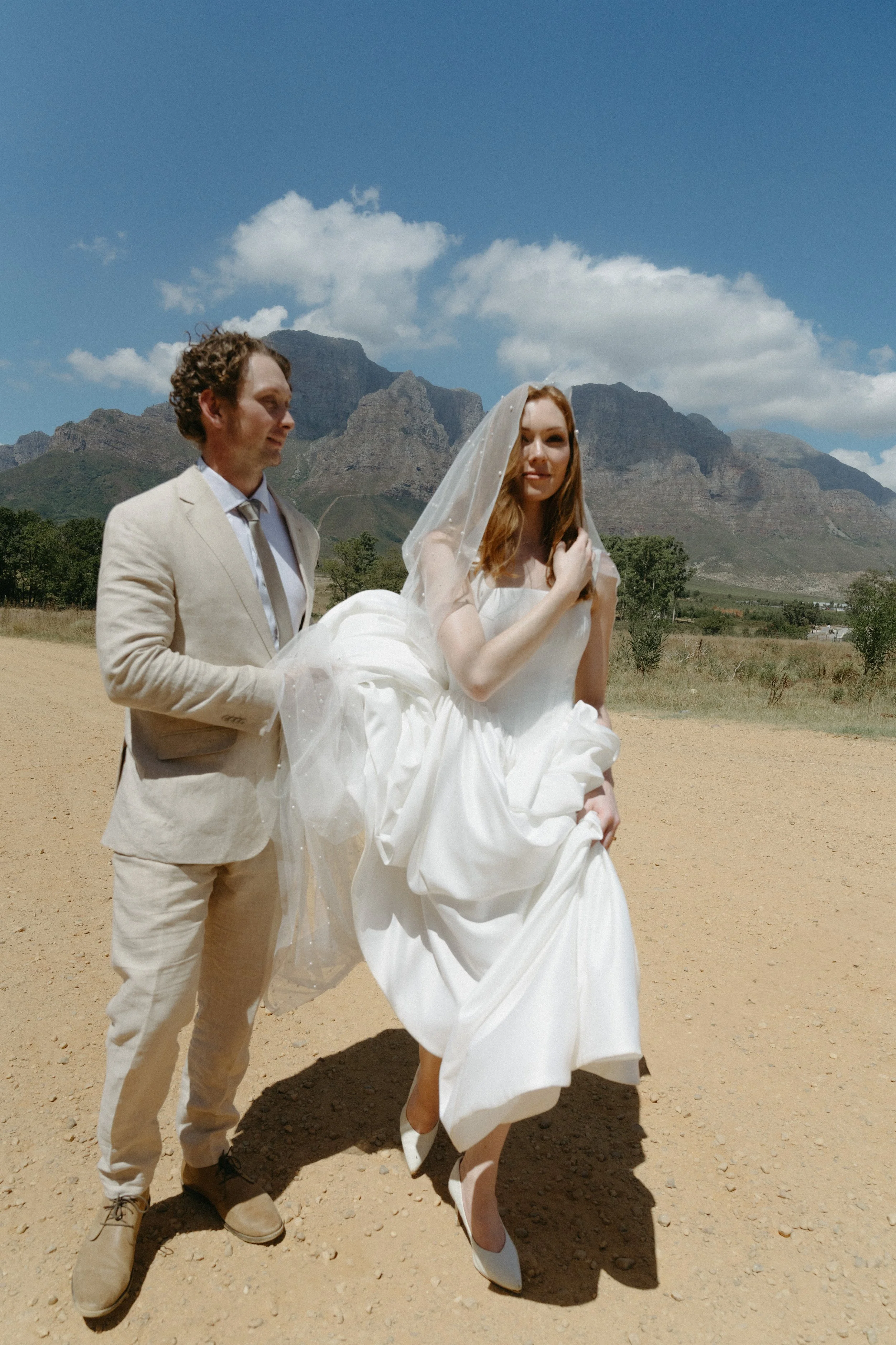 A bride in a white wedding dress and veil is being carried by a groom in a light-colored suit in an outdoor setting with mountains in the background.